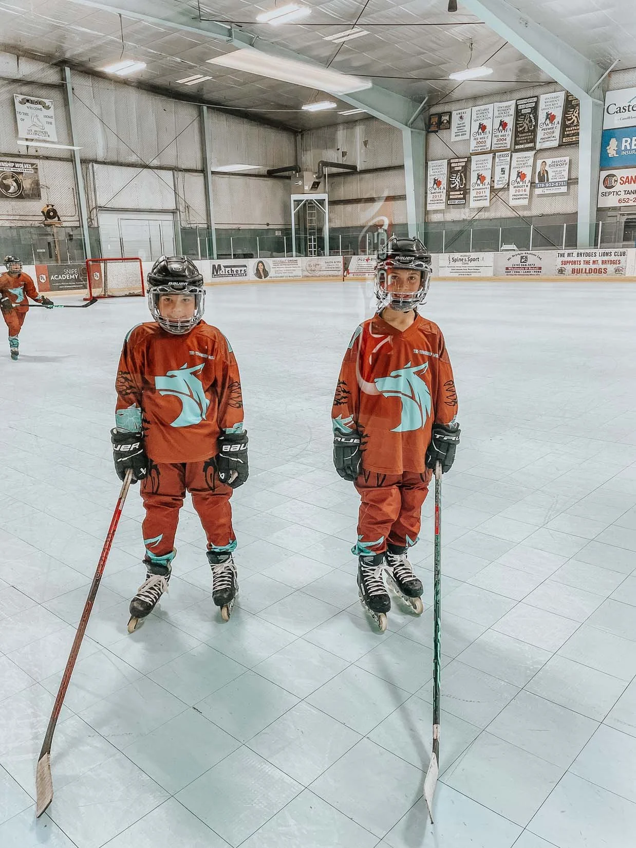 Two young boys in hockey gear standing on an ice rink, posing for a photo, with hockey sticks in their hands and wearing orange jerseys with a blue emblem, helmets, and skates.