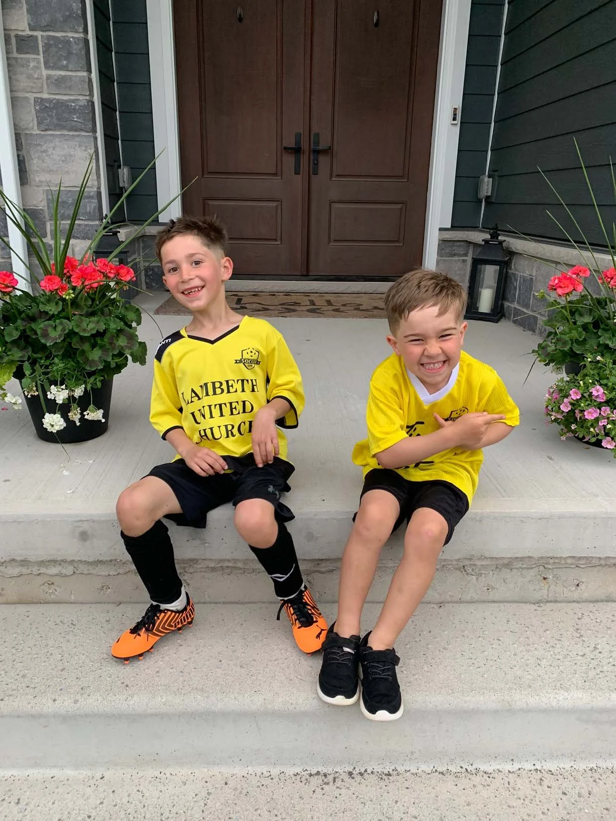 Two young boys in yellow soccer jerseys sitting on a front porch step, smiling. There are potted flowers on either side, and a wooden front door in the background.