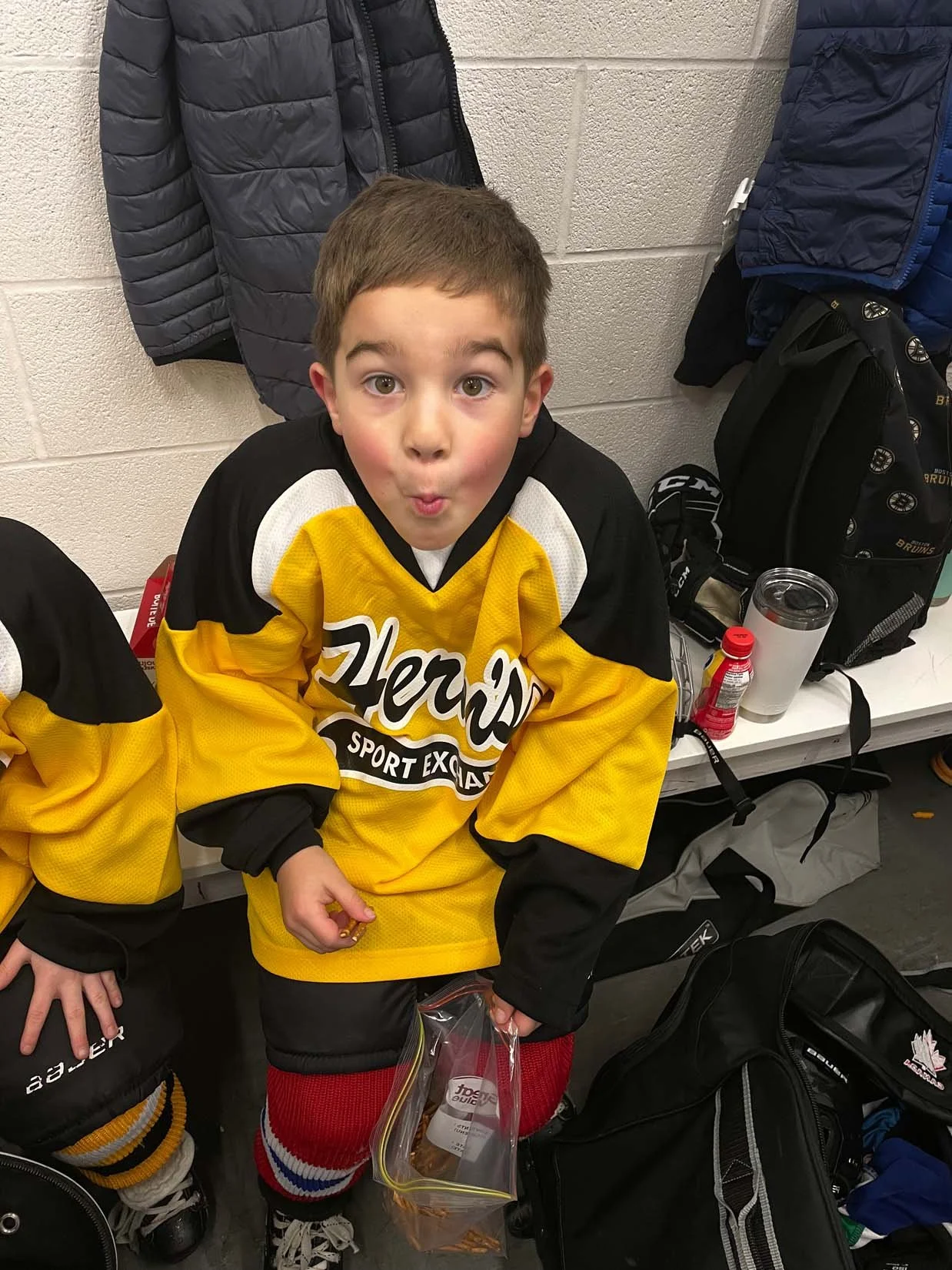 A young boy in a yellow hockey jersey making a surprised face while sitting on the floor of a locker room. He is holding a small plastic bag with snacks, and there are sports bags, jackets, and a water bottle around him.