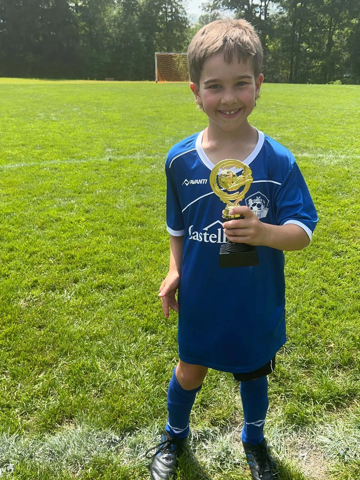 A young boy standing on a grassy field holding a trophy, wearing a blue soccer uniform and smiling after a soccer game.