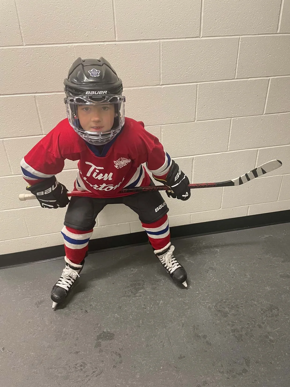 Young boy in hockey gear wearing a helmet, red hockey jersey, black shorts, striped socks, and skates, holding a hockey stick, standing on indoor ice rink floor against beige brick wall.