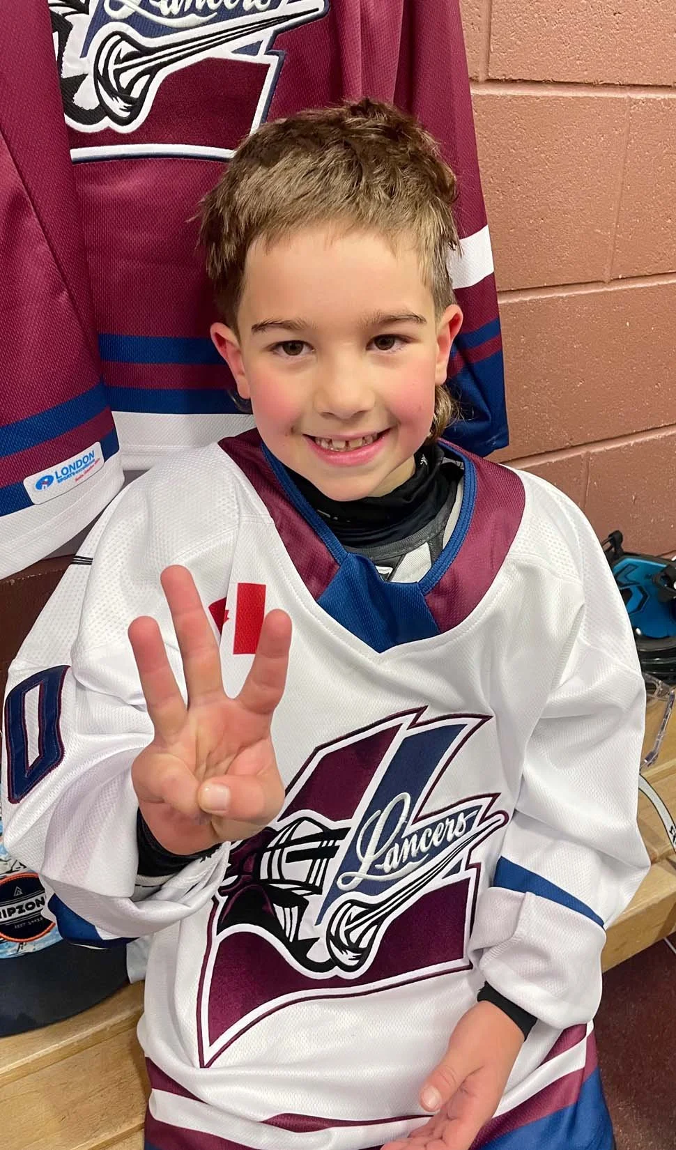 A young boy in a hockey jersey sitting on a bench, smiling, holding up three fingers in a peace sign, with jerseys and jackets hanging on the wall behind him.