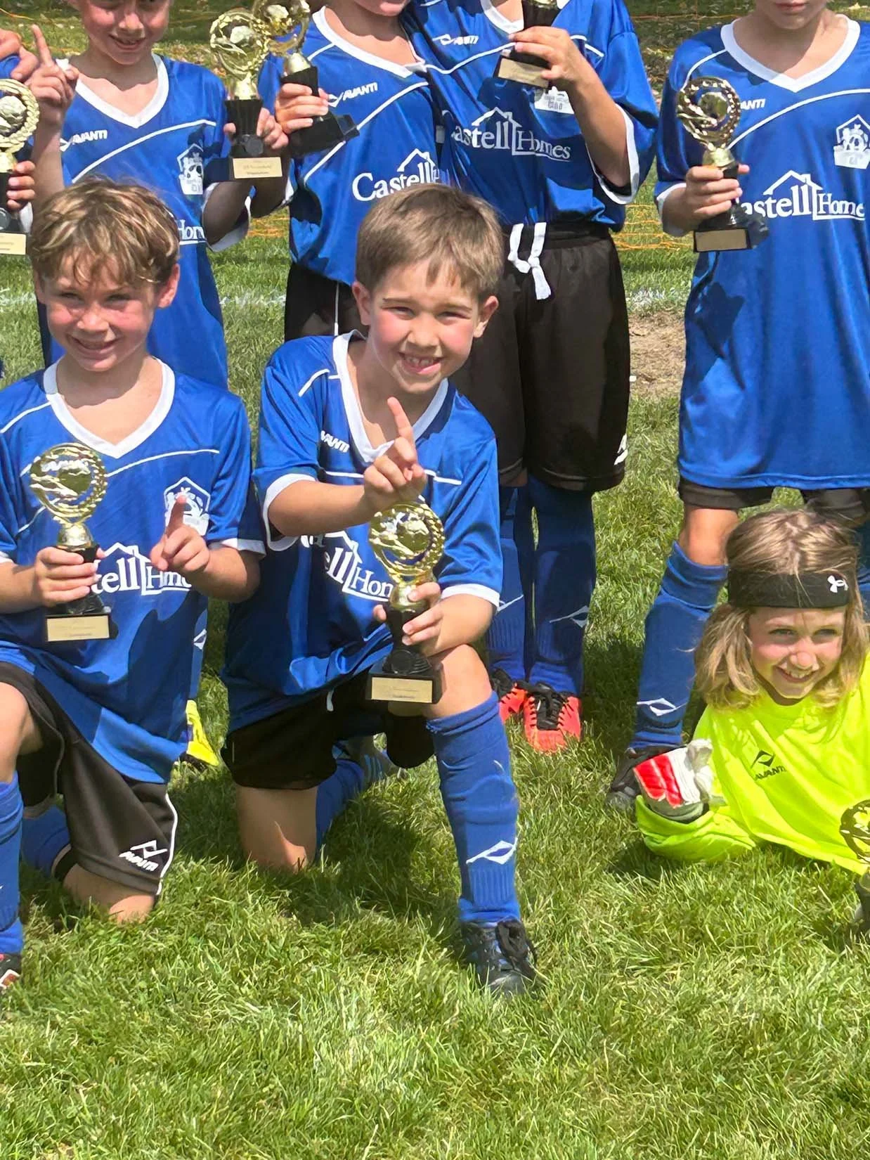 Children in blue soccer uniforms holding trophies pose for a photo on a grassy field after winning a game, with one girl in a yellow goalkeeper's jersey kneeling in front.