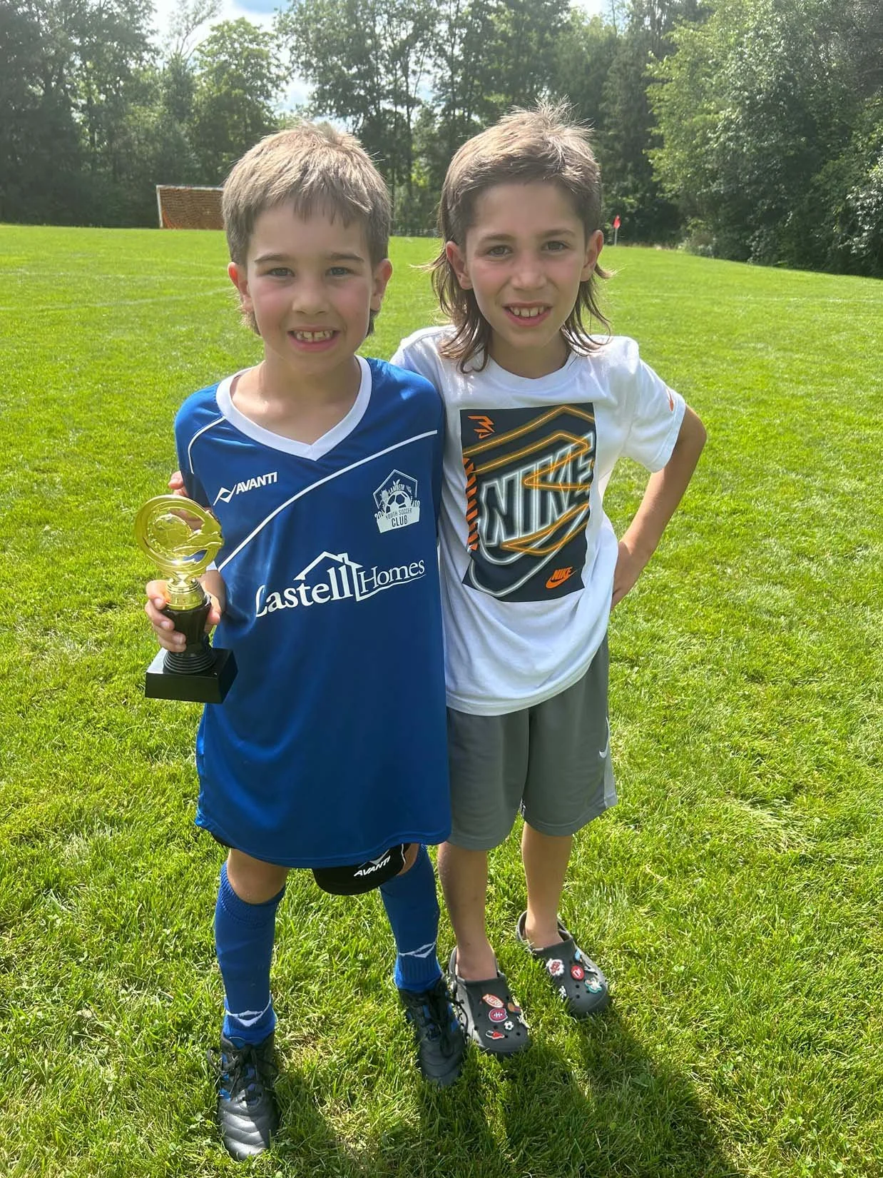 Two young boys standing on a grassy field, with one holding a small trophy, smiling at the camera, and both dressed in sports attire on a sunny day with trees in the background.