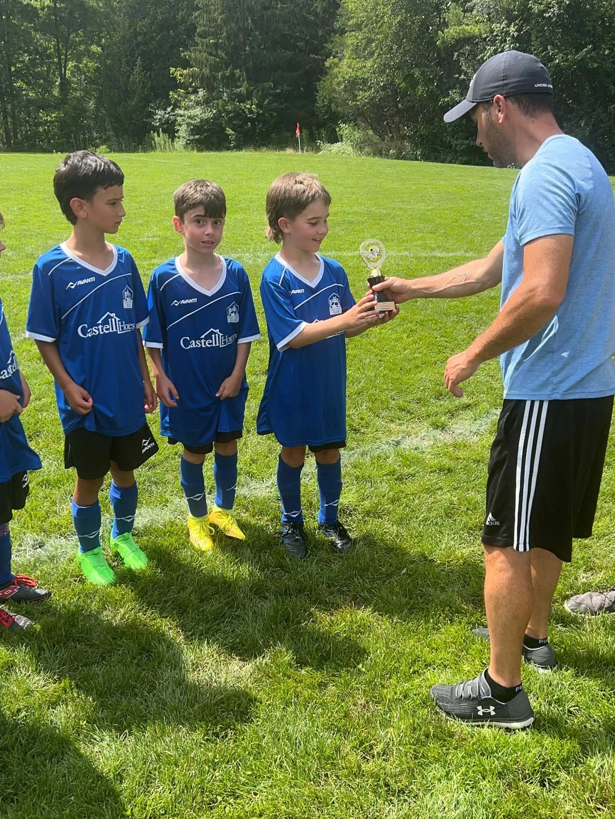 A coach presents a trophy to a young soccer player during an award ceremony on a green field, with other kids in blue soccer uniforms watching.
