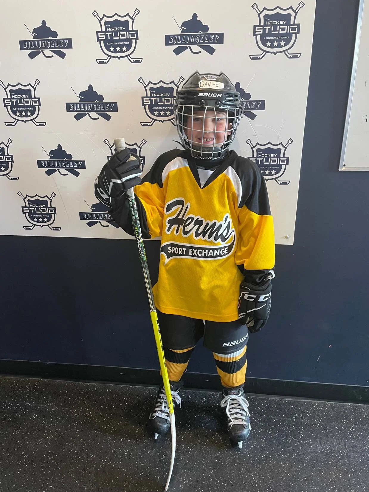 A young hockey player in black and yellow uniform with the logo 'Herm's Sport Exchange', wearing a helmet, gloves, and skates, standing on indoor ice hockey rink, holding a hockey stick, smiling with a thumbs-up gesture. Behind him is a wall with pos