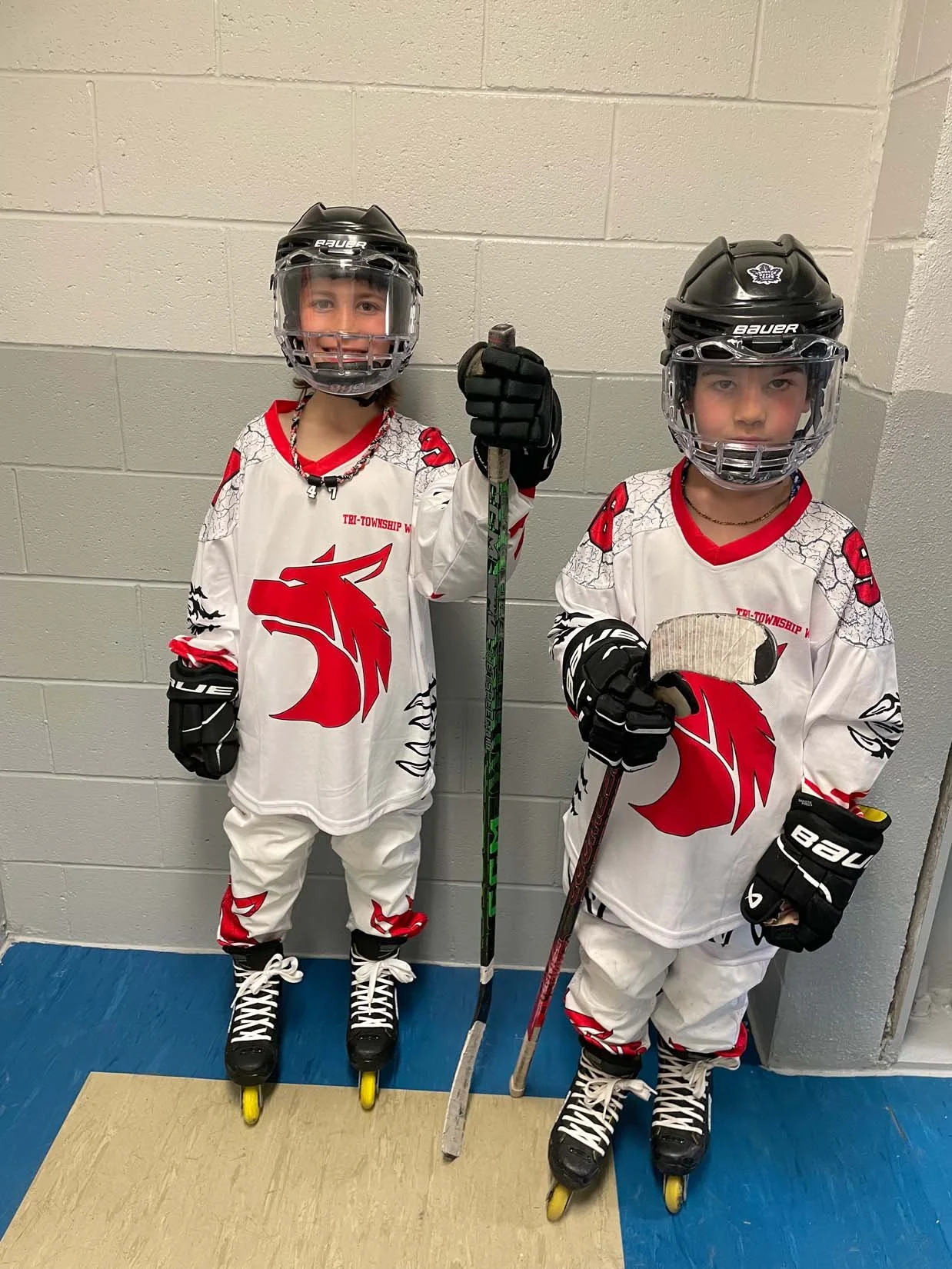Two young boys wearing hockey gear, including helmets, gloves, skates, and jerseys with a red lion logo, posing with hockey sticks against a gray cinder block wall.
