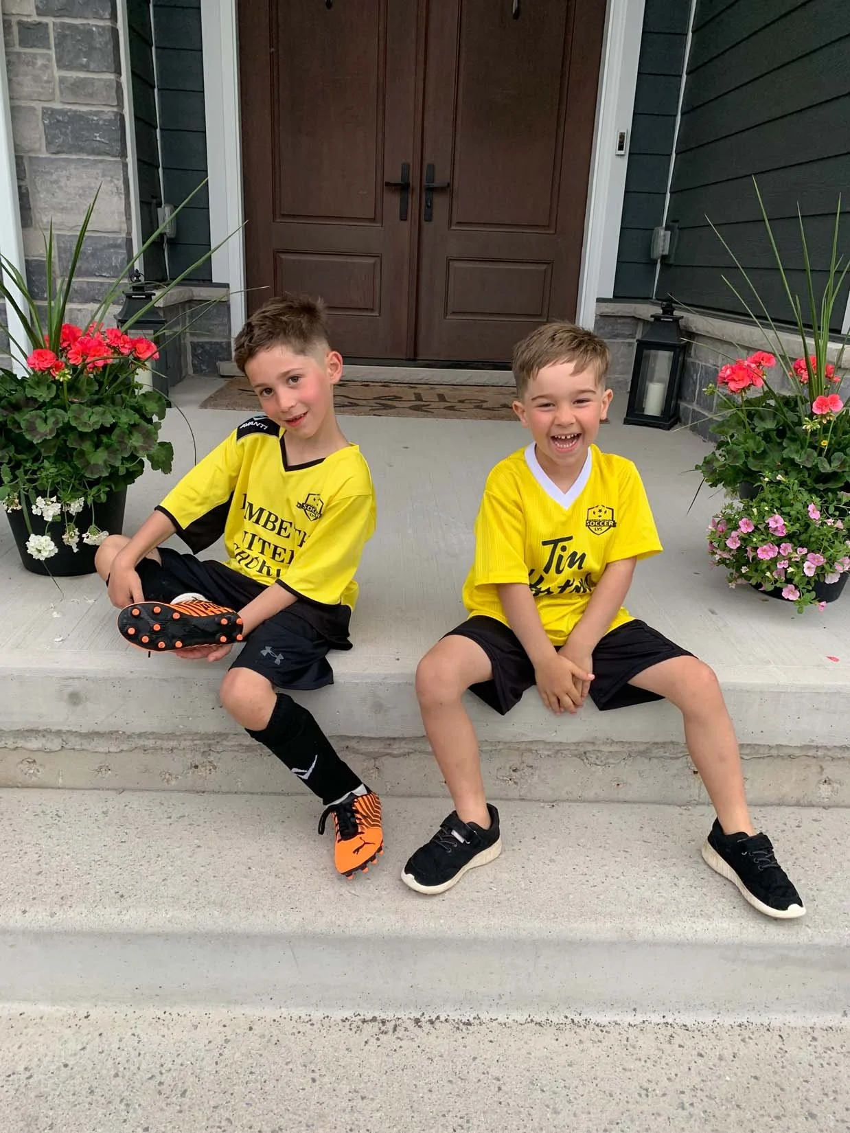 Two young boys sitting on a front step, wearing yellow soccer jerseys, smiling. There are potted flowers on either side of them, and a wooden front door behind.
