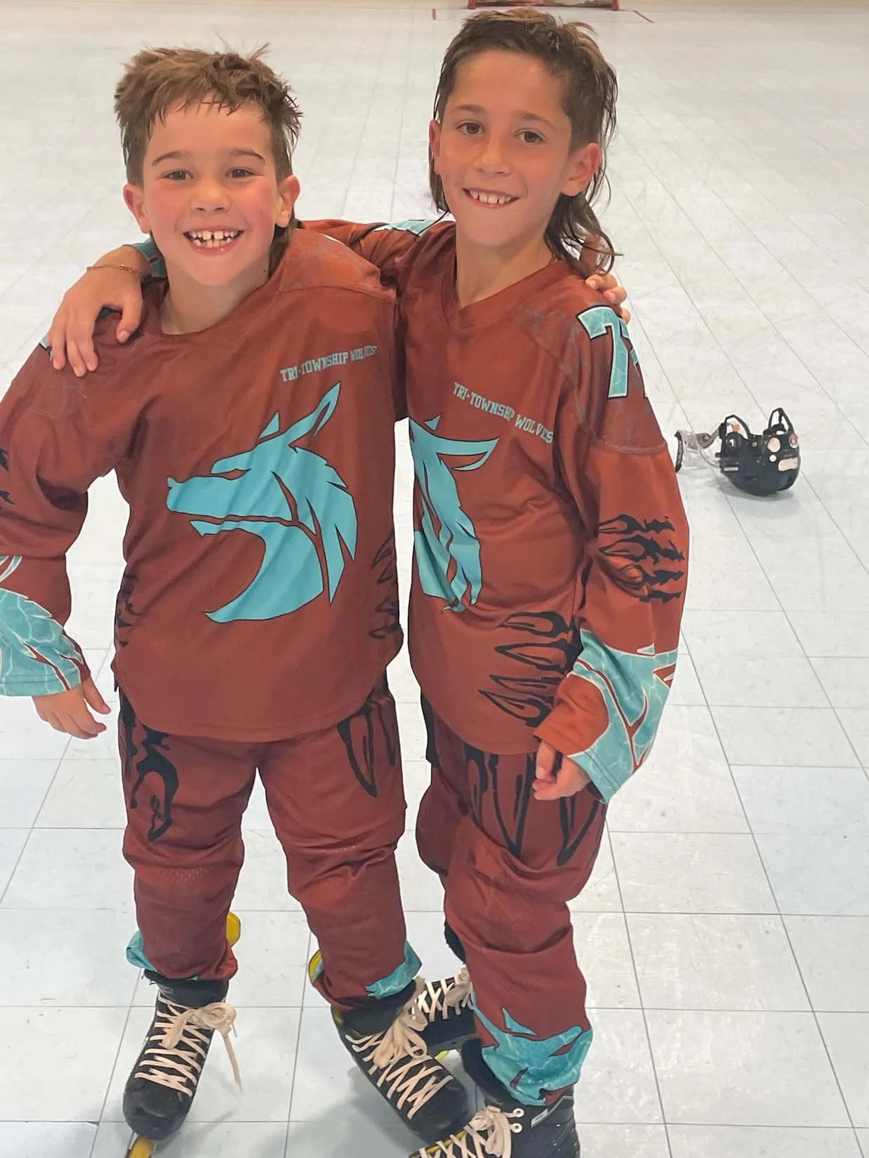 Two young boys wearing matching sports jerseys and rollerblades, smiling and standing together on an indoor rink.