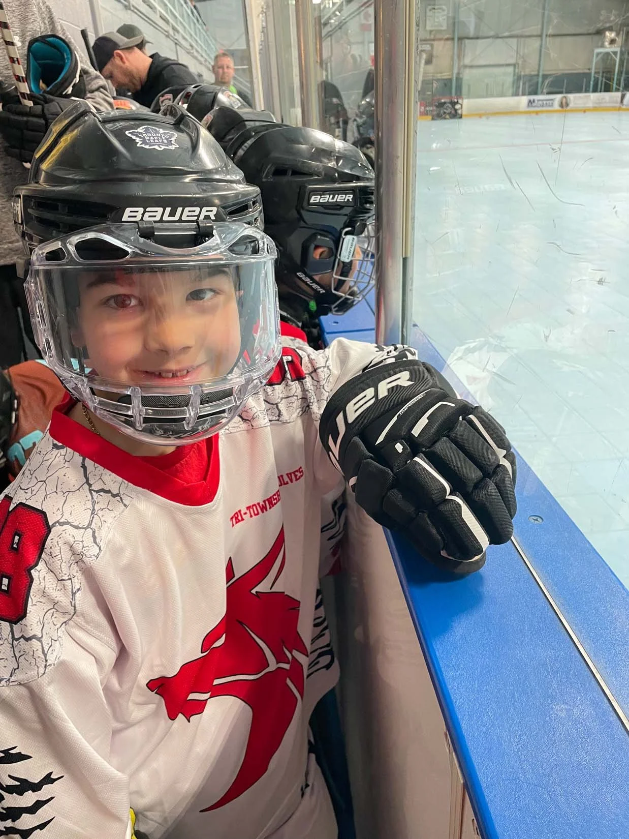 Young boy dressed in hockey gear, including a helmet with a clear face shield, hockey gloves, and a white jersey with a red lion logo, sitting at the edge of an ice rink inside the hockey arena. He is smiling and looking at the camera. Behind him, th