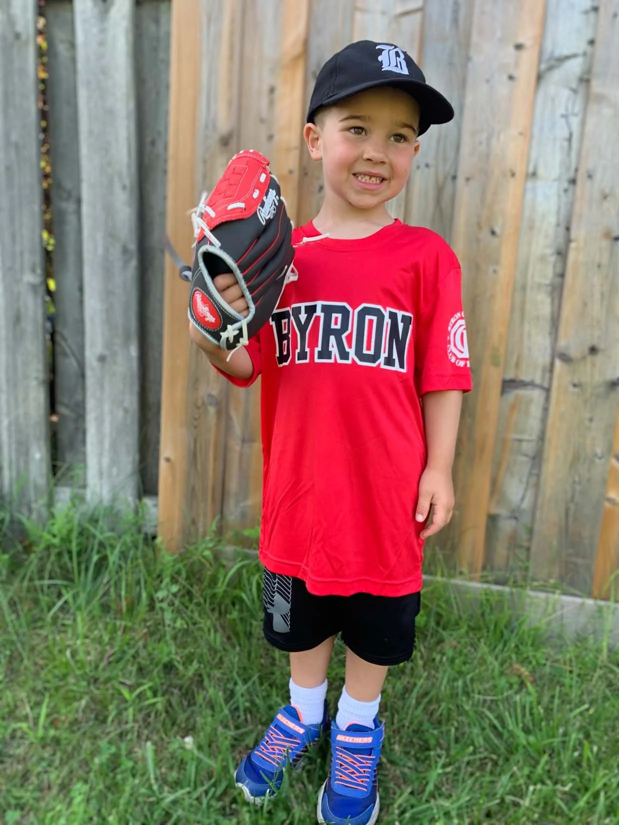 A young boy in a red Byron shirt, black shorts, and blue sneakers standing outdoors in front of a wooden fence, holding a baseball glove in his right hand, wearing a black baseball cap, and smiling.