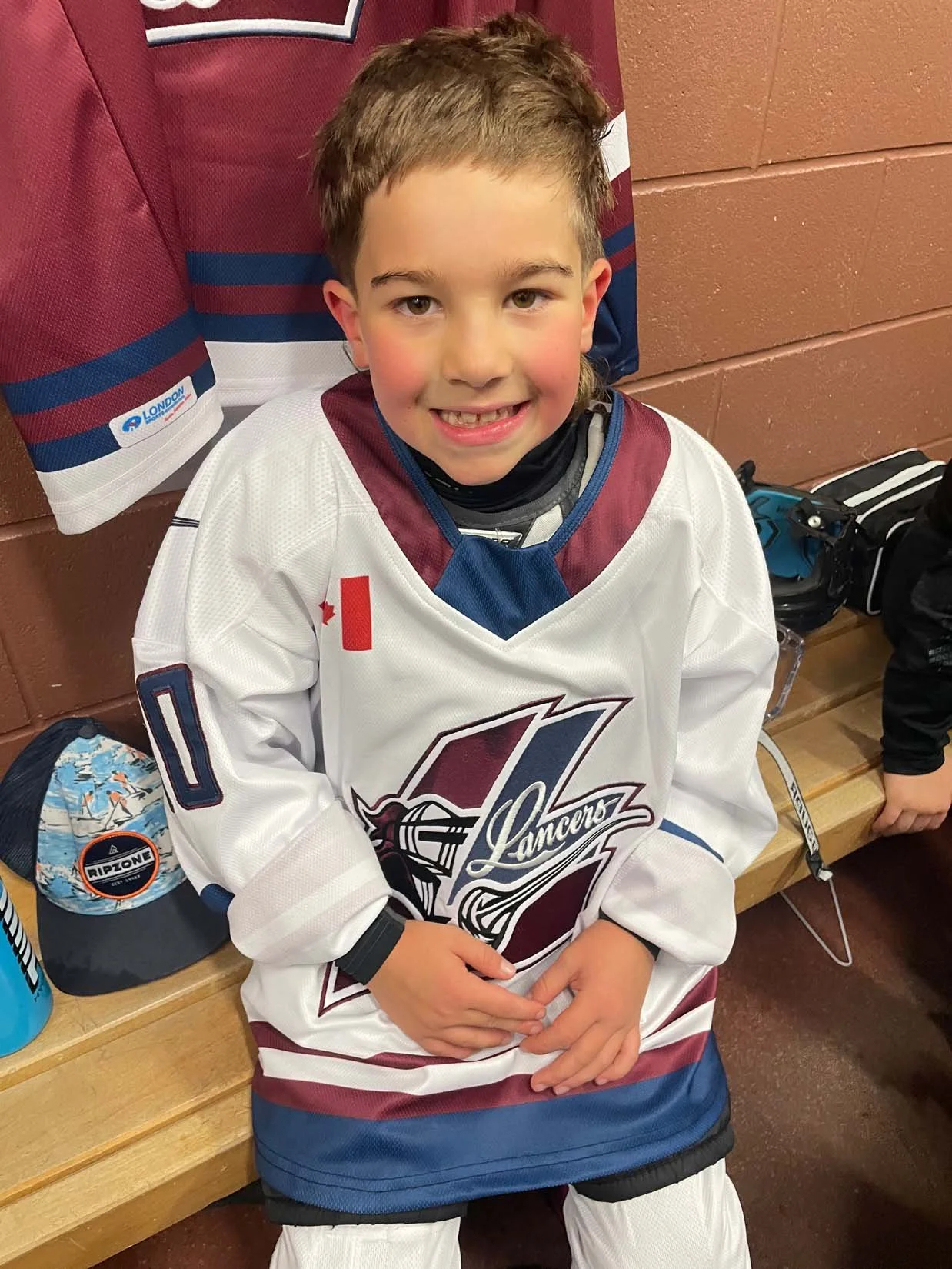 A young boy in a hockey uniform sitting on a bench, smiling at the camera. He is wearing a white hockey jersey with maroon and blue accents, and has a black neck guard and a black wristband.