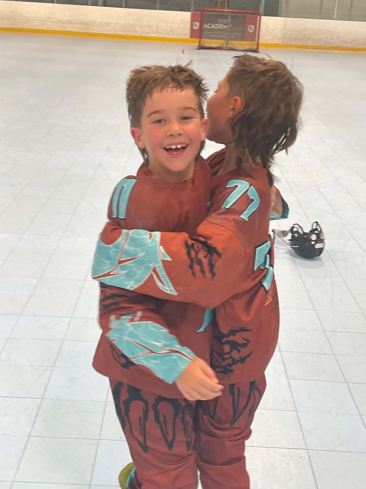 Two young boys in hockey uniforms hugging each other on an indoor ice rink, with one boy smiling at the camera.