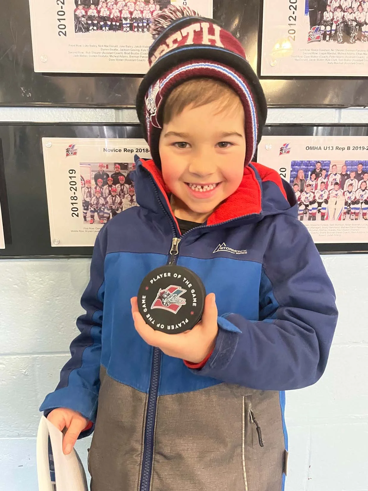 A young boy wearing a winter jacket and a red hockey hat is smiling and holding a hockey puck that says 'Player of the Game'. He is standing in front of hockey team photos on the wall.