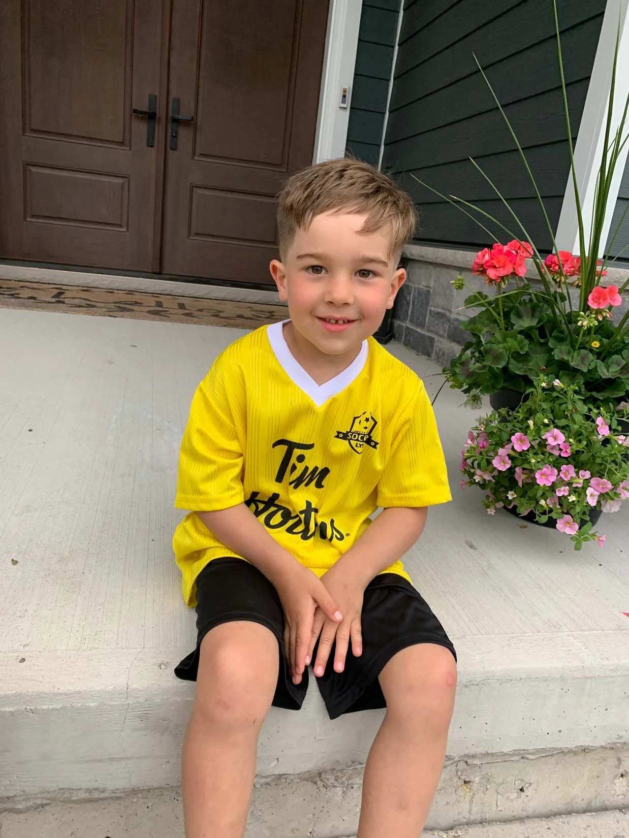 A young boy sitting on a concrete porch in front of a wooden door and a house with dark siding. He is wearing a yellow soccer jersey and black shorts, smiling at the camera with a devious look. There are pink and red flowers in a pot next to him.