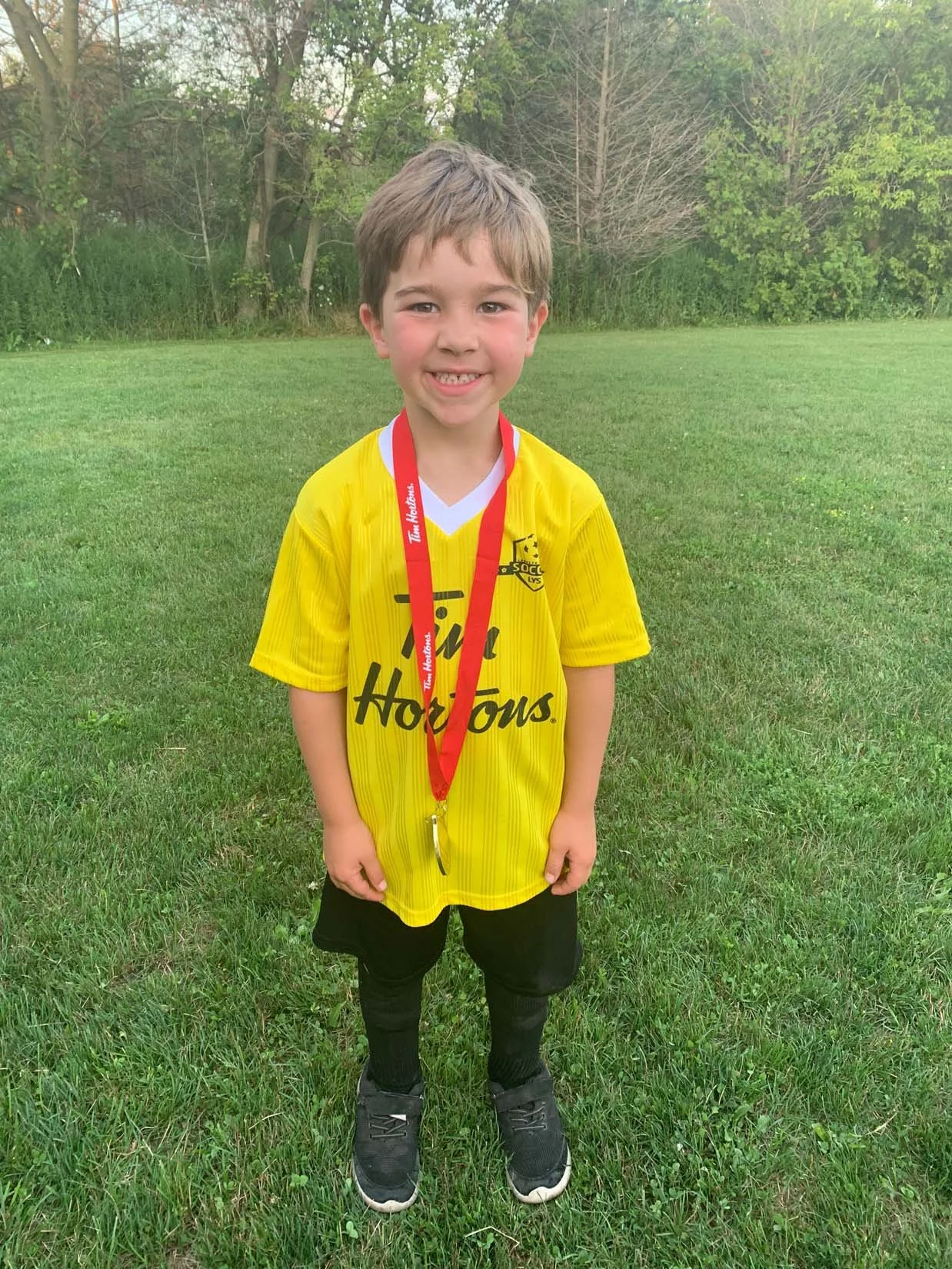 Young boy standing on a grassy field, wearing a yellow soccer jersey with the word 'Hots' on it and a red medal around his neck, smiling at the camera.