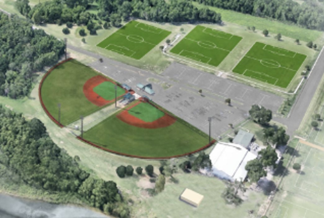 Aerial view of a sports complex with soccer fields, a baseball field, a parking lot, and surrounding trees.