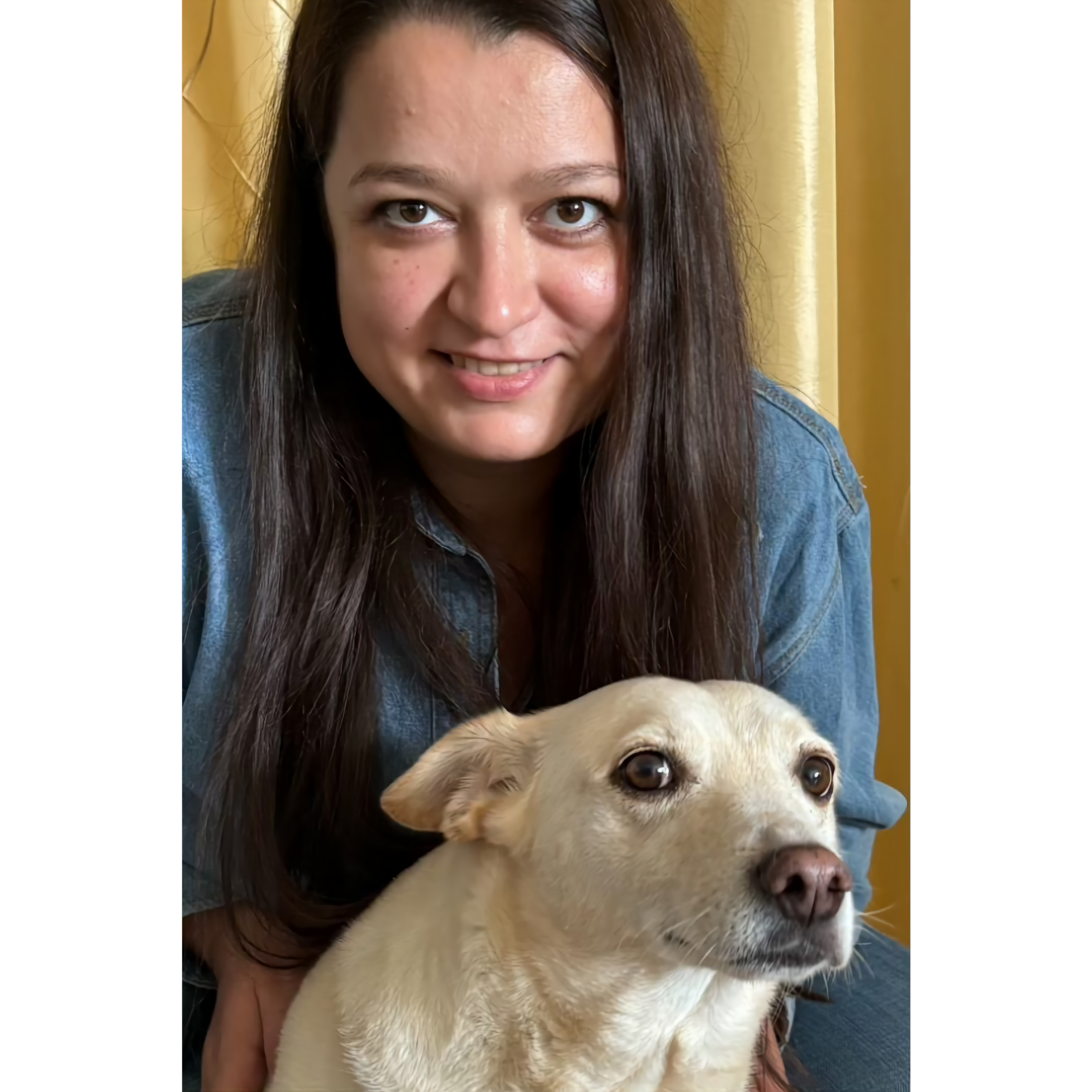 A woman with long dark hair smiling, wearing a denim shirt, leaning over a light-colored dog with brown eyes and a pink nose, indoors with yellow curtains in the background.