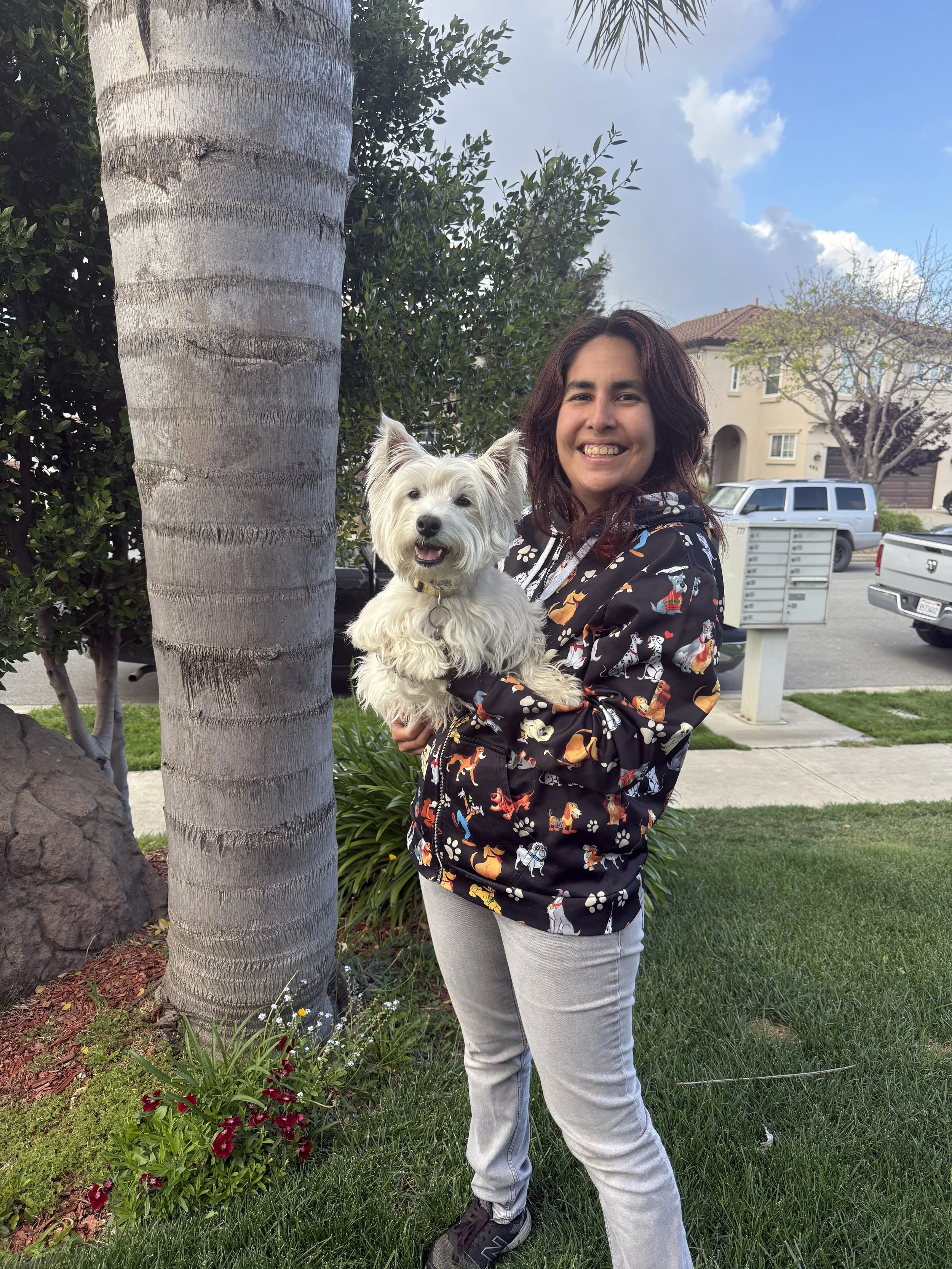 A smiling woman with dark brown, wavy hair holding a small white dog with fluffy fur outdoors near a palm tree and houses in the background.