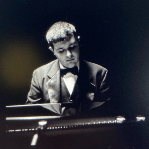 A black and white image of a young man wearing a suit and bow tie, playing the piano with a focused expression.