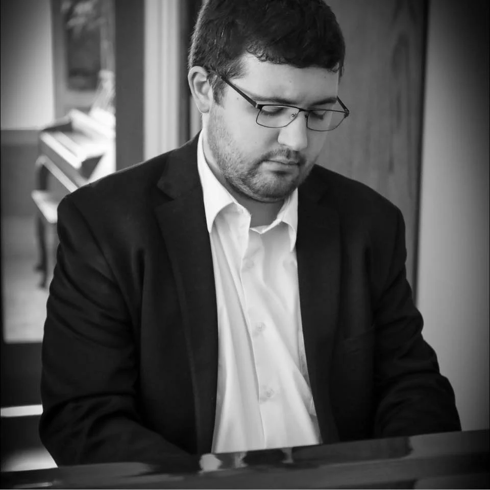 A man with glasses and dark hair, wearing a suit and white shirt, sitting at a piano with a focused expression.