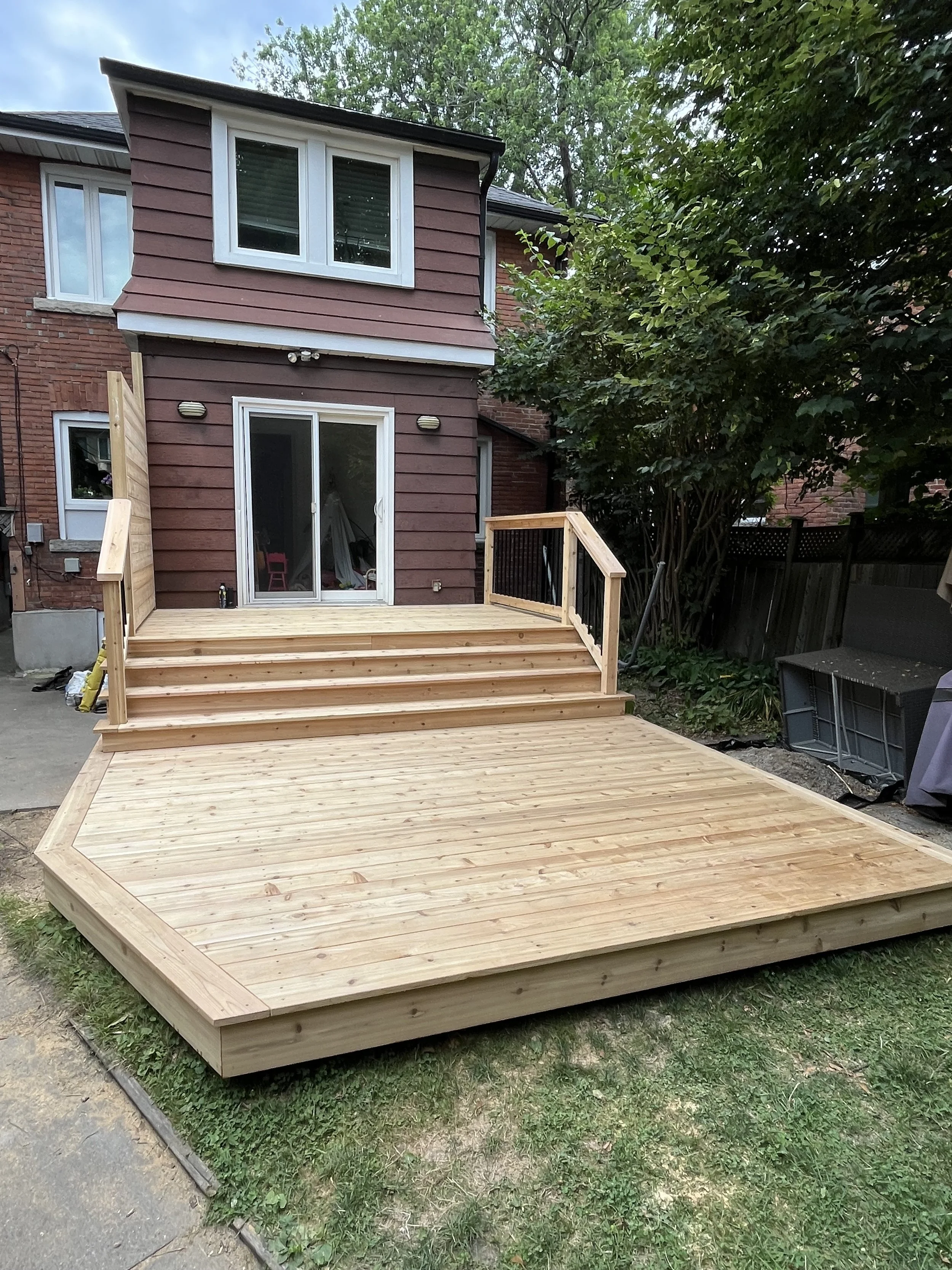 Newly built wooden deck with stairs attached to the back of a red brick house, surrounded by trees and a fence.