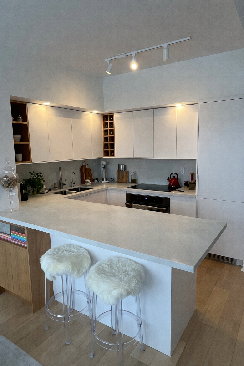 Modern kitchen with white cabinets, a white countertop, and two transparent bar stools with fur cushions. The countertop has various tools and items, and the kitchen is well-lit with ceiling spotlights.