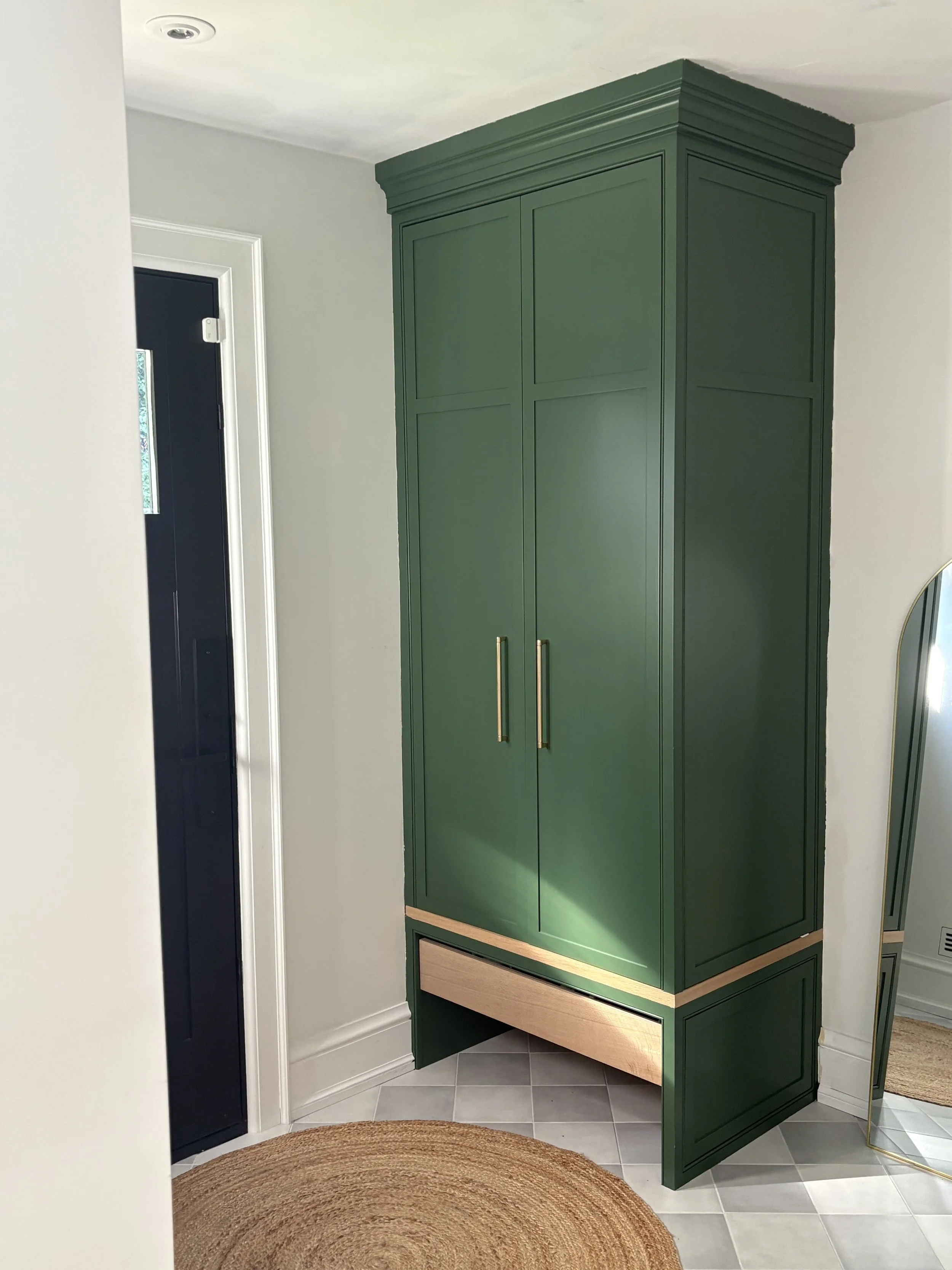 Green corner cabinet with gold handles in a hallway, next to a door and a mirror, with a circular woven rug on tiled floor.