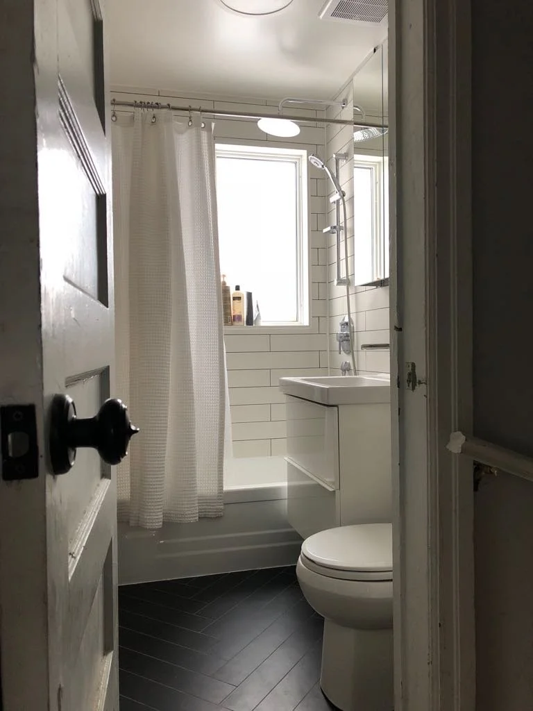 A small modern bathroom with a white bathtub and shower, white tiled walls, a window, a white sink with a cabinet, a toilet, and a dark wood floor viewed through an open door.