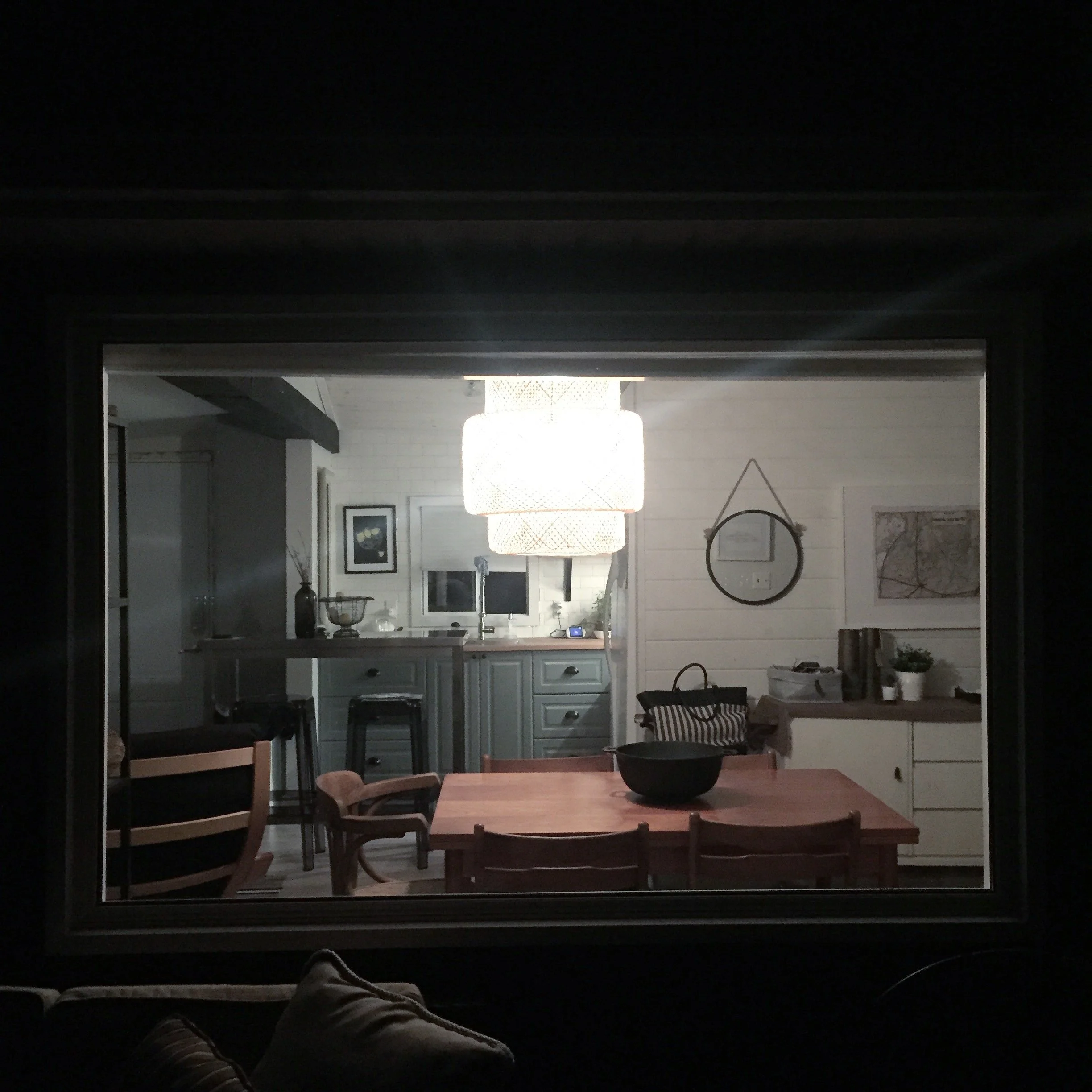 View of a cozy, modern kitchen and dining area through a window at night, with a large ceiling light, wooden dining table, chairs, and decorative wall art.
