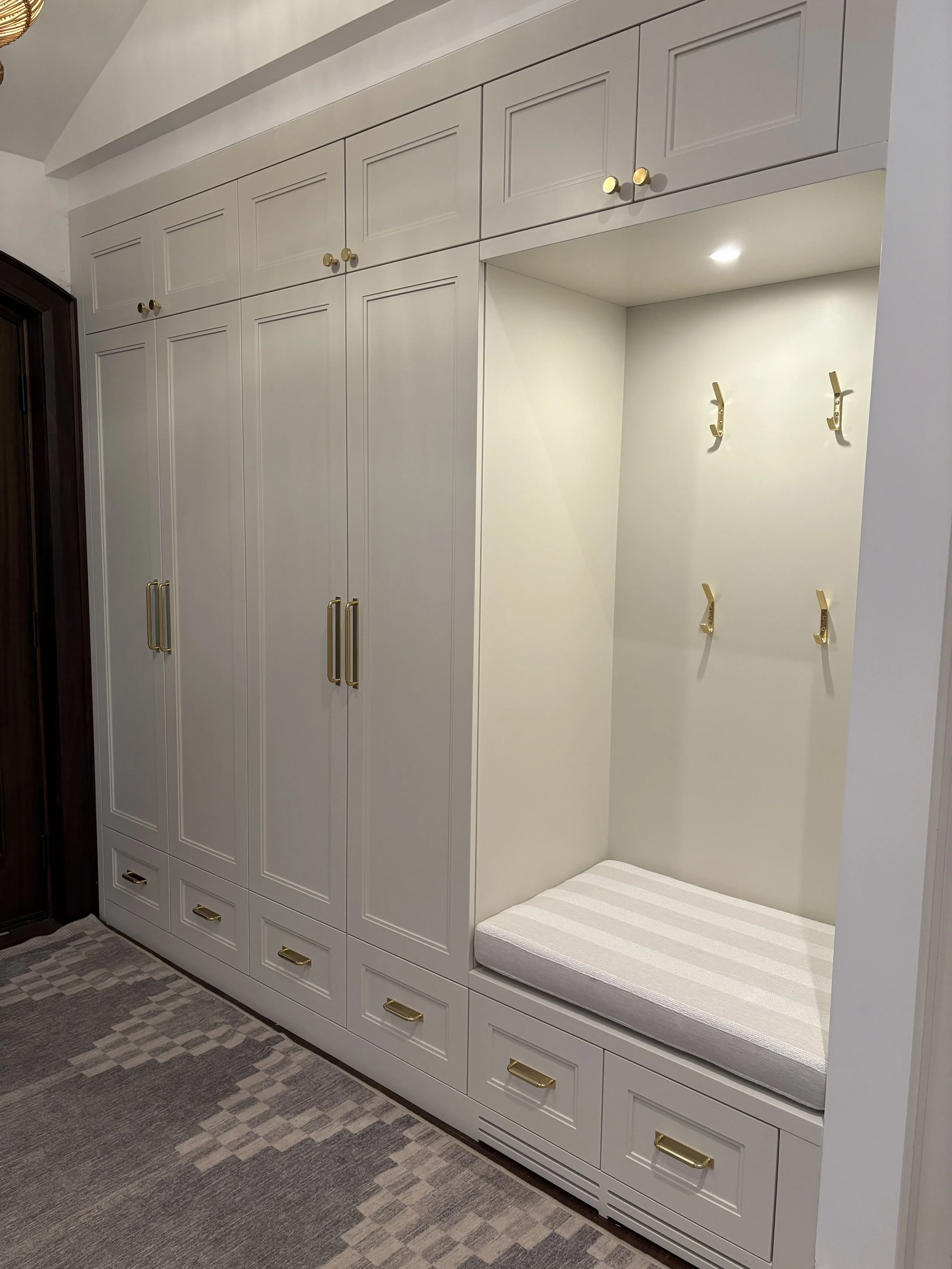 White built-in closet with gold handles, hooks, and a cushioned seating area, located in a modern room with patterned gray carpet.