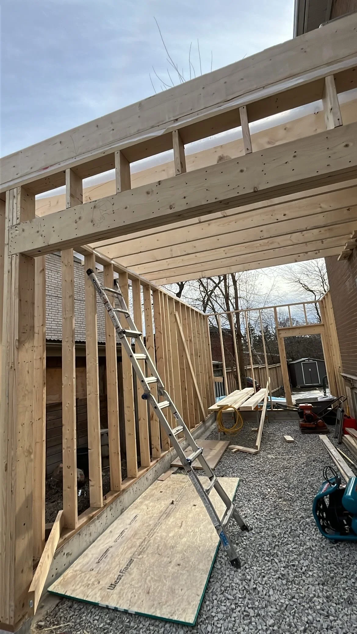 Construction of a wooden outdoor structure with framing, ladder, and building materials on gravel ground.