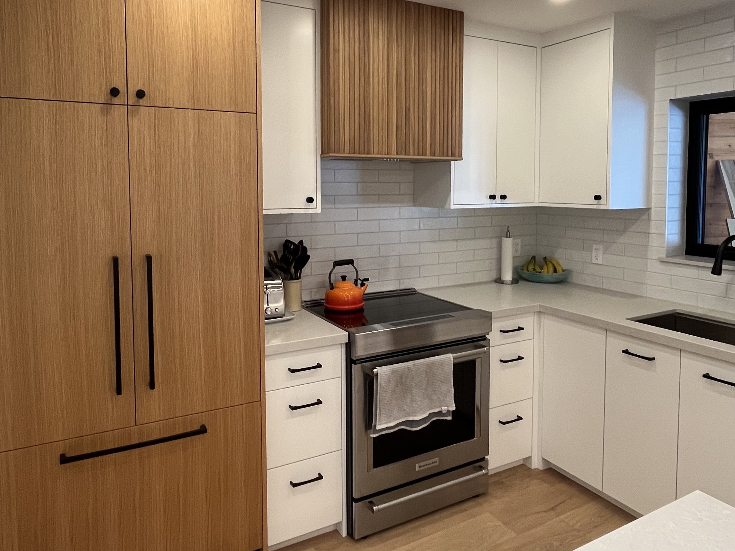 Modern kitchen with white cabinets, black handles, a stainless steel oven, a black faucet, a window, a paper towel holder, a bowl of bananas, and wooden flooring.