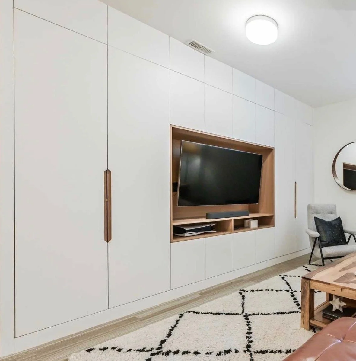 Living room with a large white built-in wall unit, a mounted flat-screen TV, a grey armchair with a black pillow, a wooden coffee table, and a patterned rug.