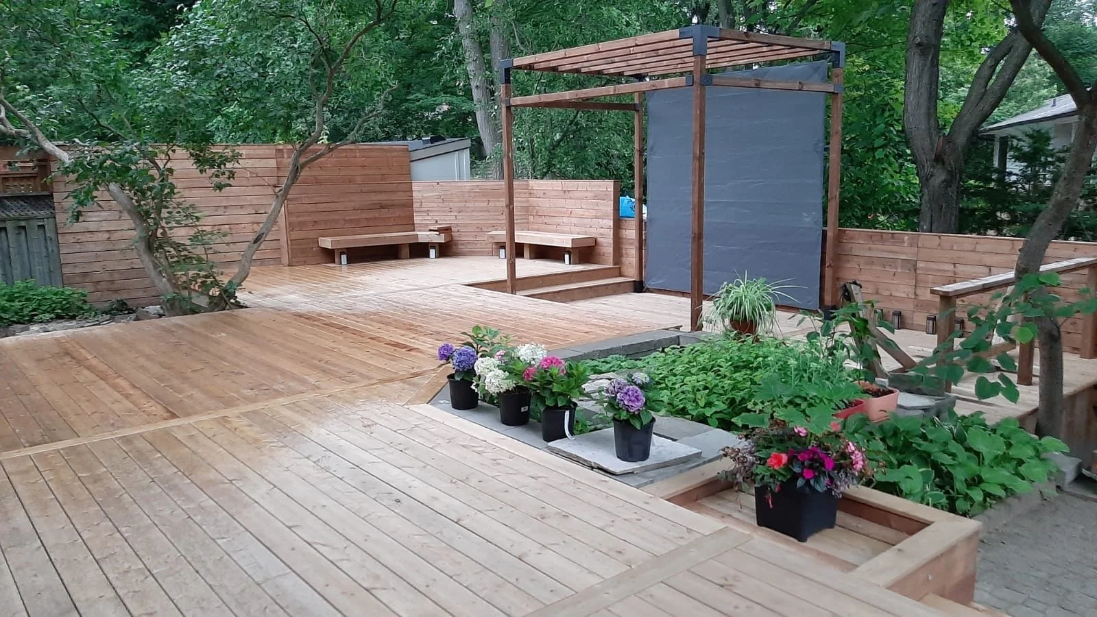 A backyard deck with wooden flooring, a small garden with potted plants, flower beds, built-in benches, trees, and a privacy fence.