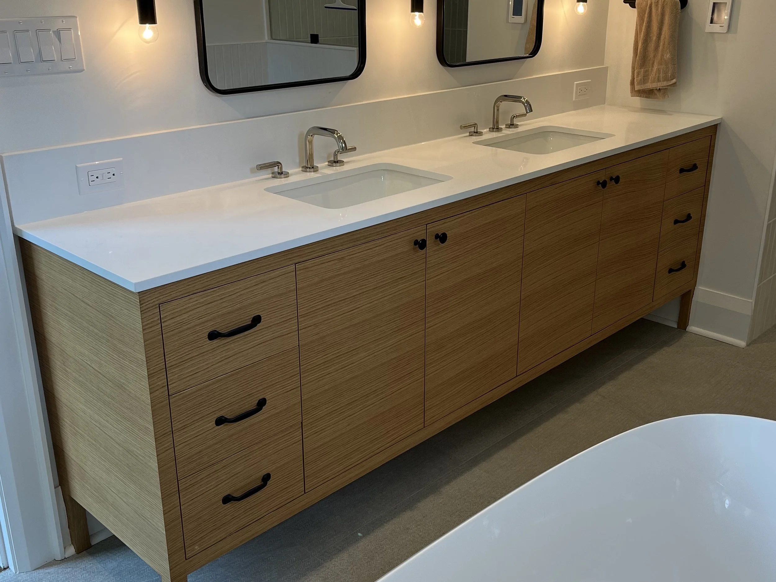 A modern bathroom vanity with a white countertop, two sinks, and threecentred mirrors. Wooden cabinets with black handles, and a round bathtub partially visible in the foreground.