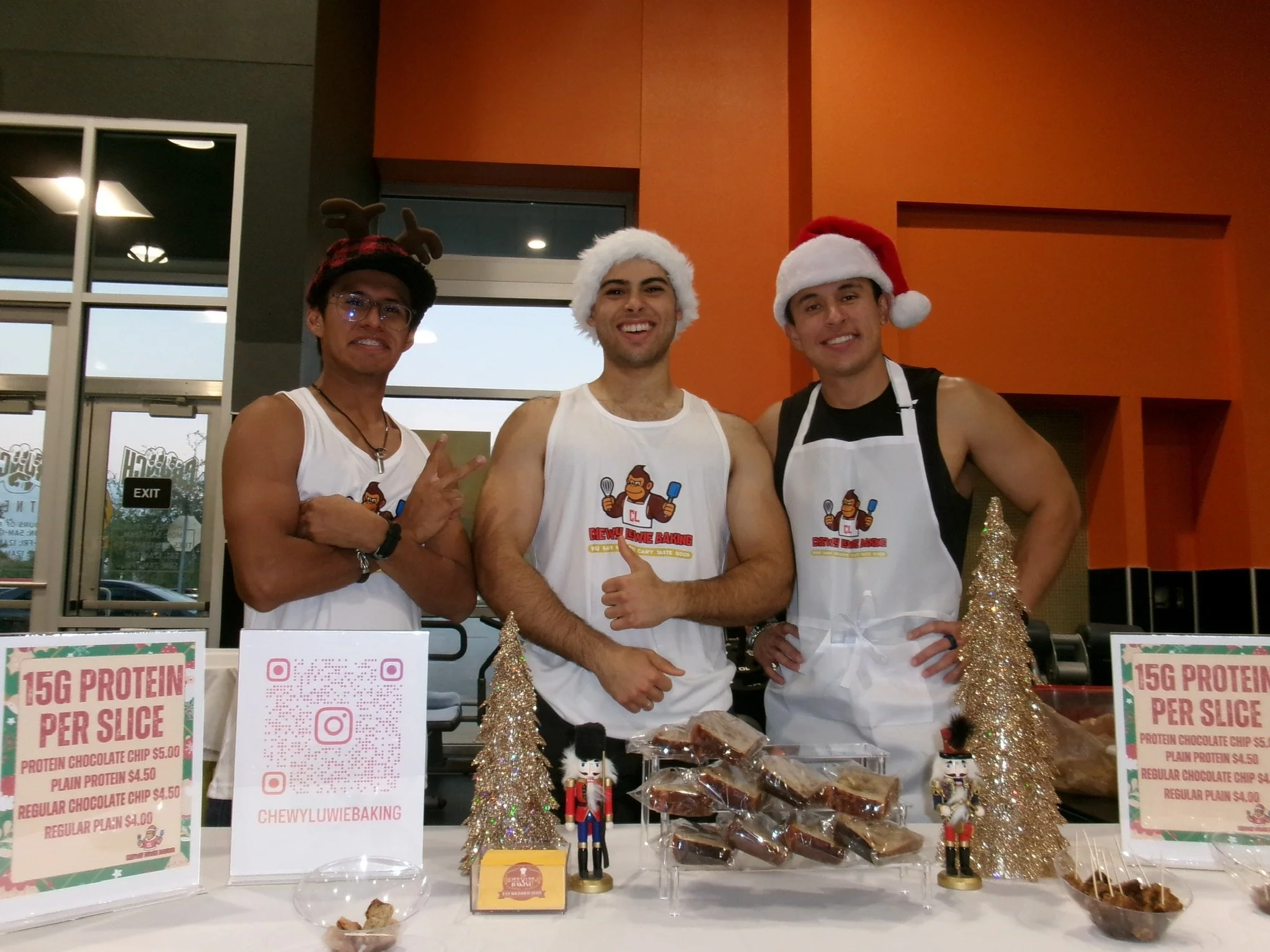 Three men wearing Christmas hats and holiday-themed clothing standing behind a table with baked goods at a holiday market or event.