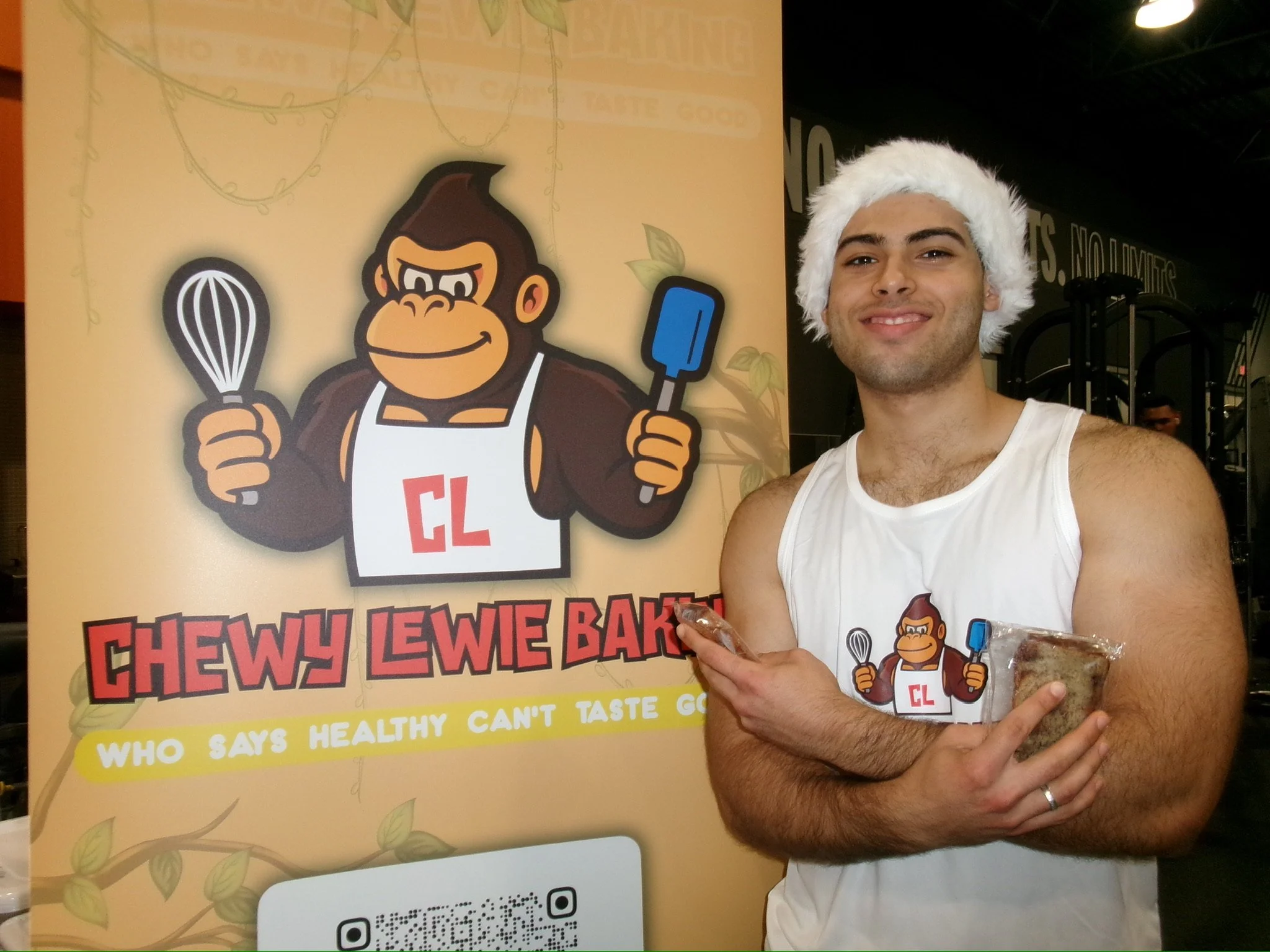 A young man with a Santa hat, wearing a Chewy Lewie Bake shirt, holding a packaged baked good. He is standing next to a Chewy Lewie Bake promotional sign featuring a cartoon ape holding a whisk and a spatula.