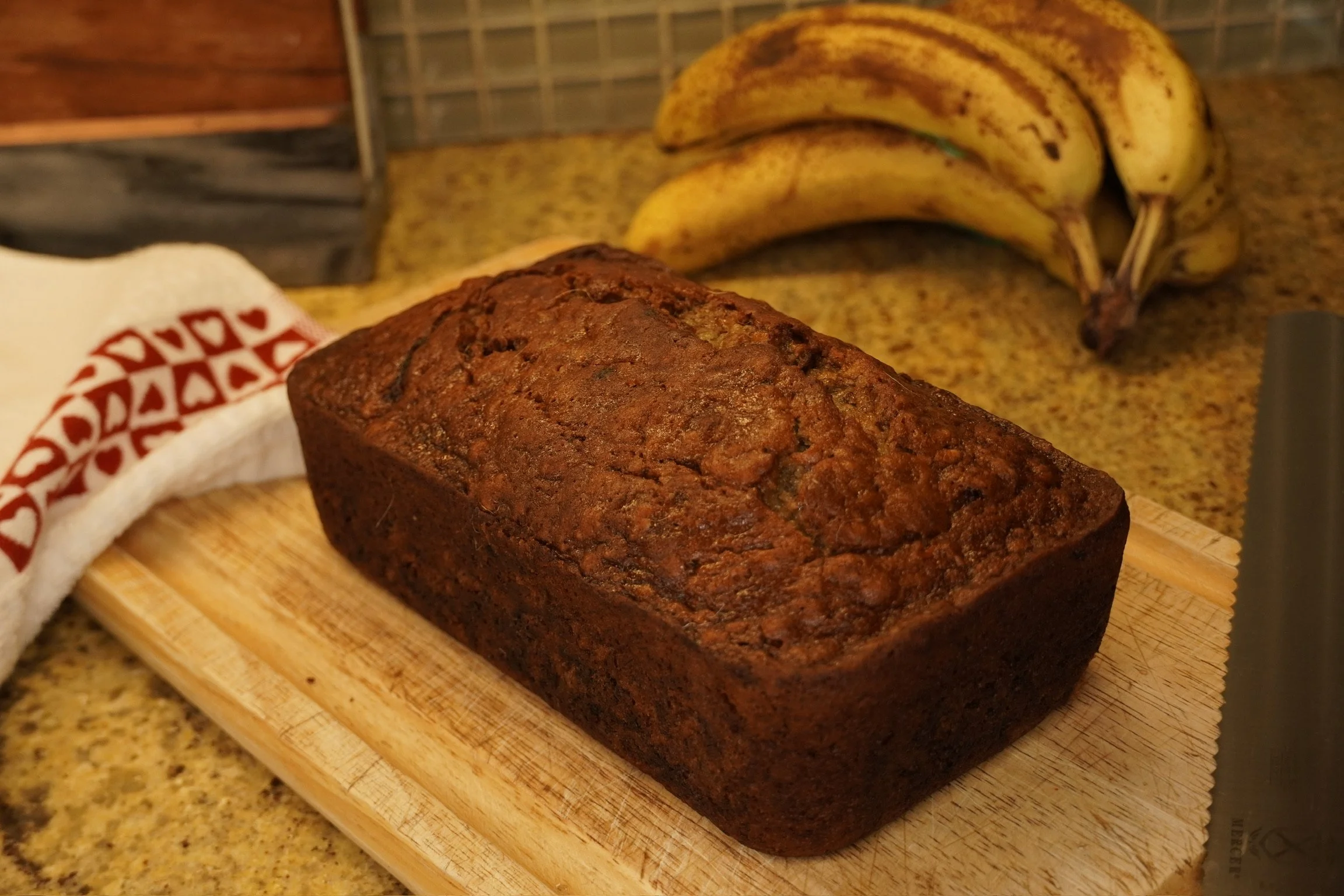 A loaf of freshly baked banana bread on a wooden cutting board, with a bunch of ripe bananas and a chef's towel in the background.
