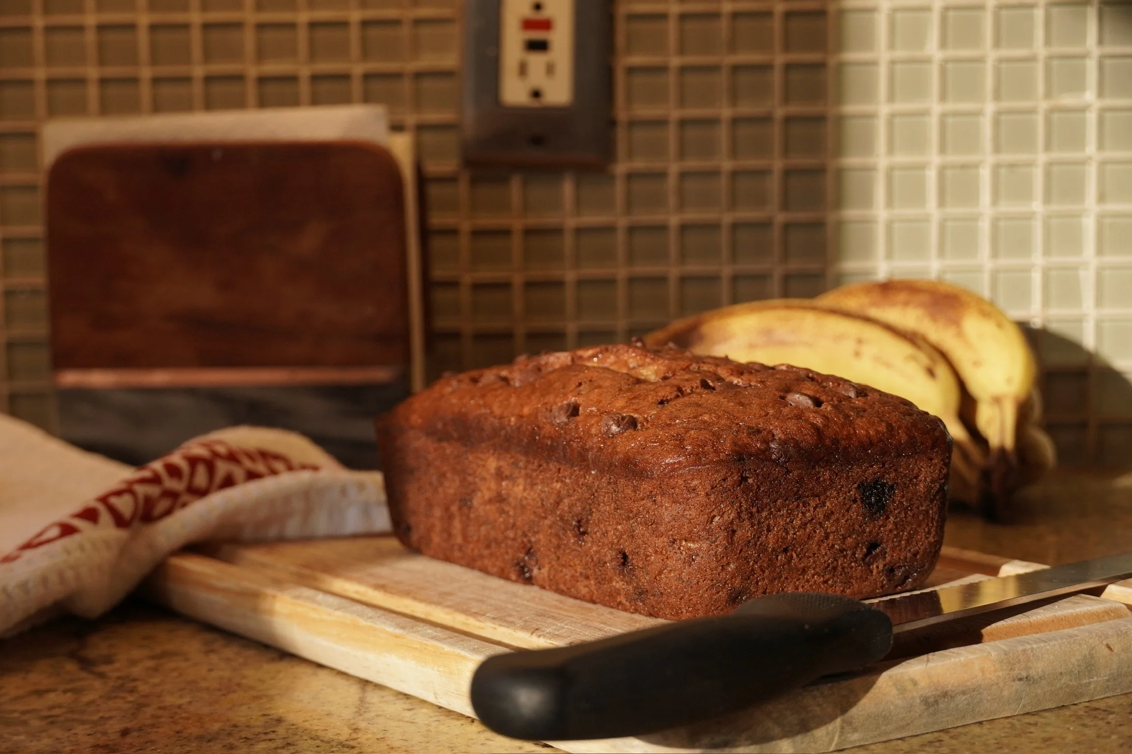 Loaf of banana bread on a wooden cutting board with a knife, bananas in the background, and a vintage wall clock.