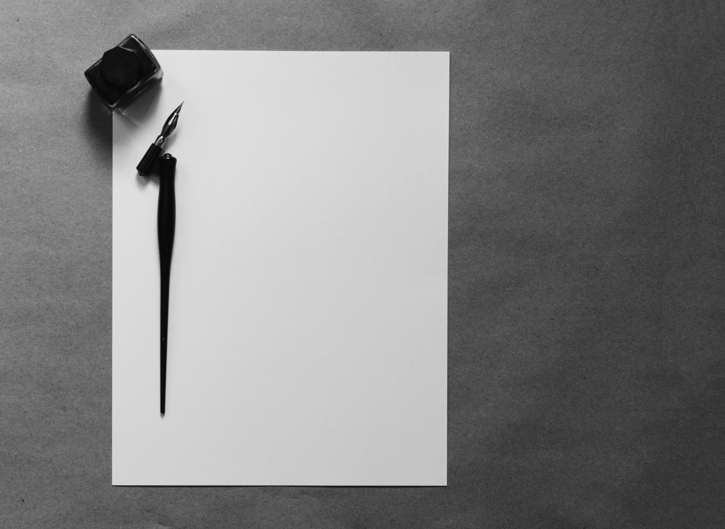 A blank sheet of white paper, a black fountain pen with its cap off, and a black ink bottle on a gray textured background, arranged neatly.