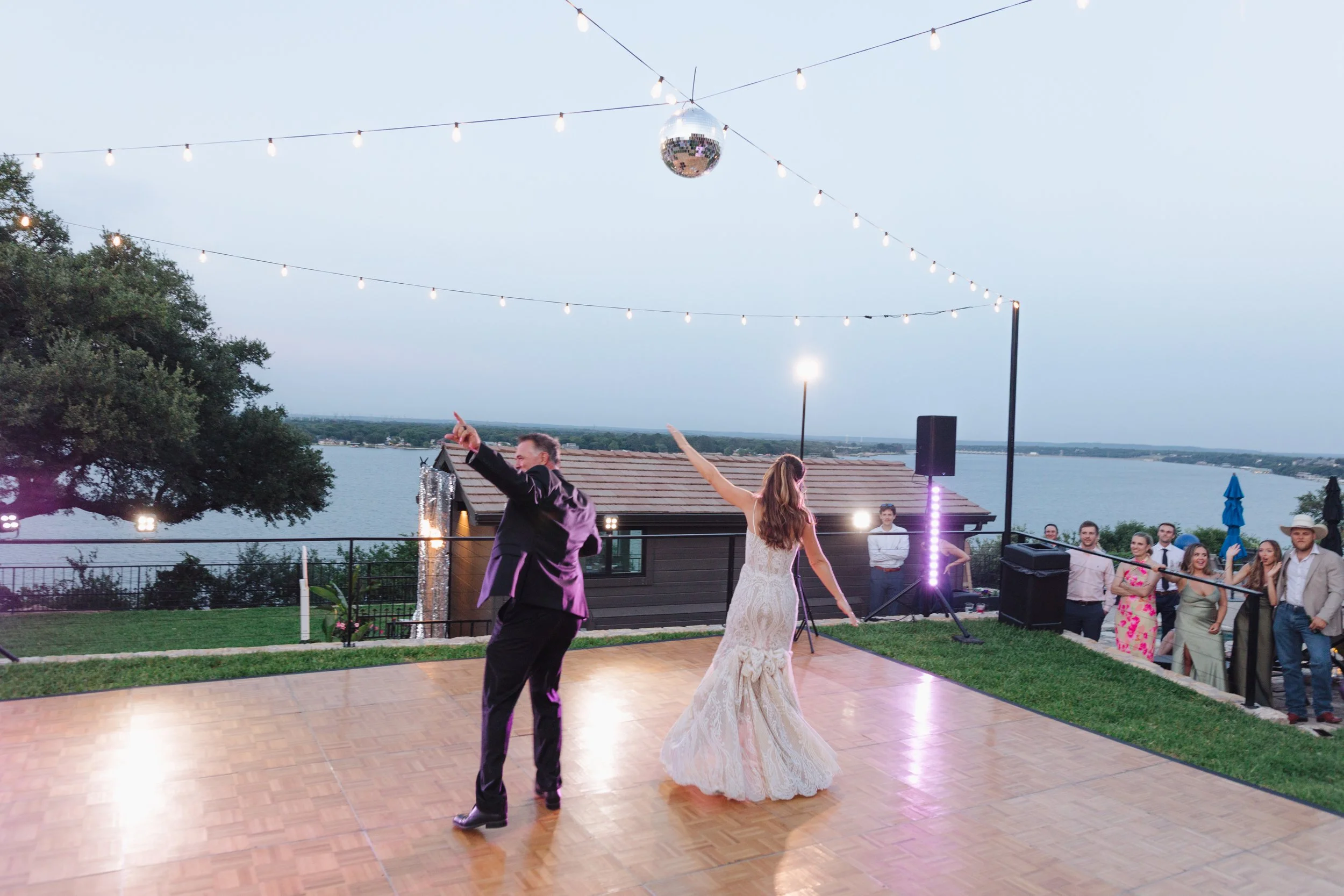 A bride and groom dancing on an outdoor wooden dance floor at sunset with string lights overhead, with guests watching nearby and a lake in the background.