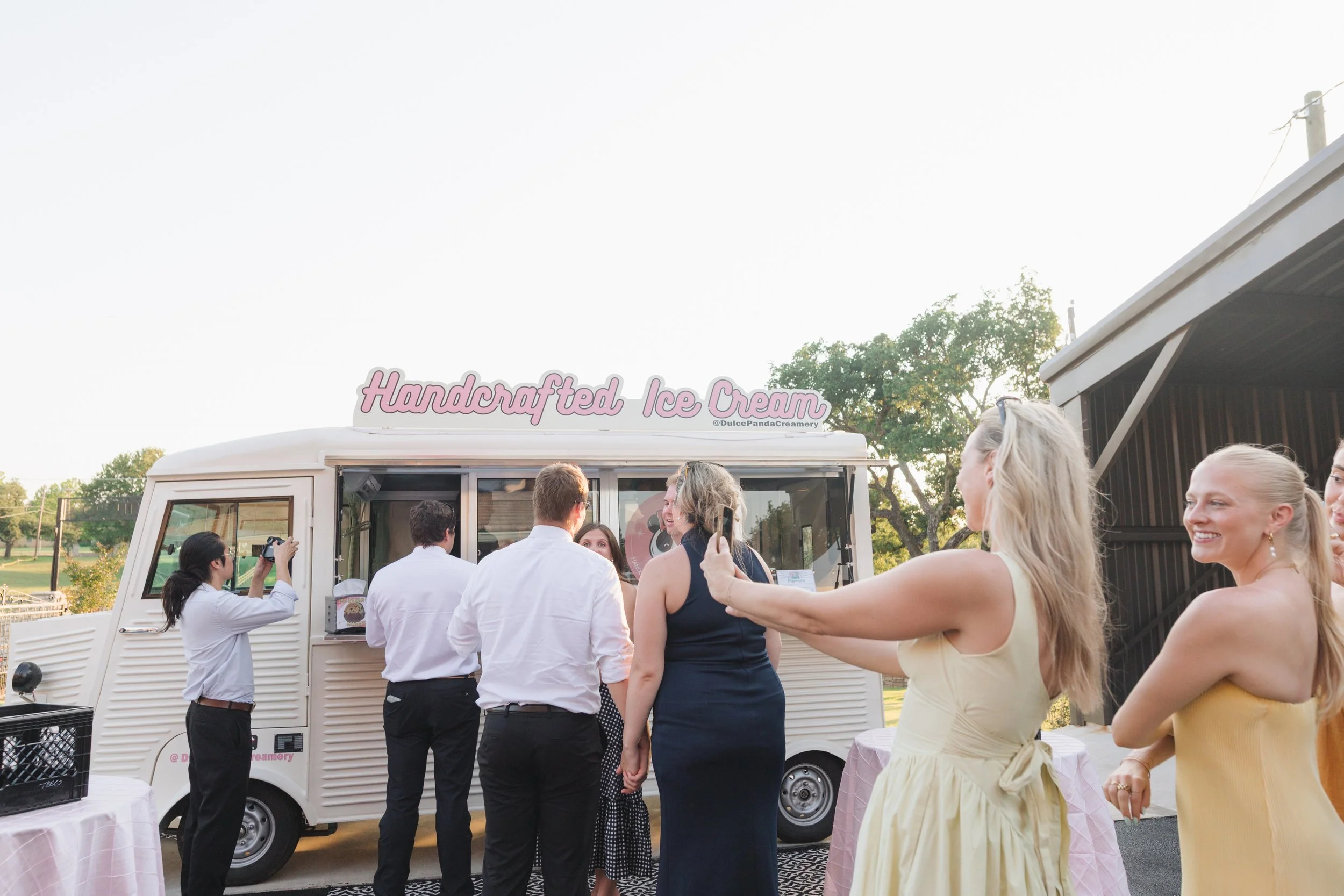 People gathered around a white ice cream truck with a sign that reads 'Handcrafted Ice Cream'. Some are taking photos and smiling, outdoors during daytime.