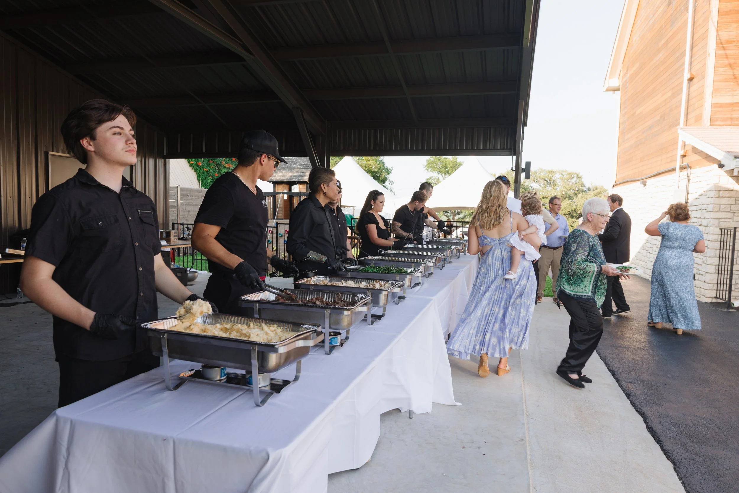 People attending an outdoor buffet with food serving stations, some serving themselves and others waiting, under a covered area during daytime.