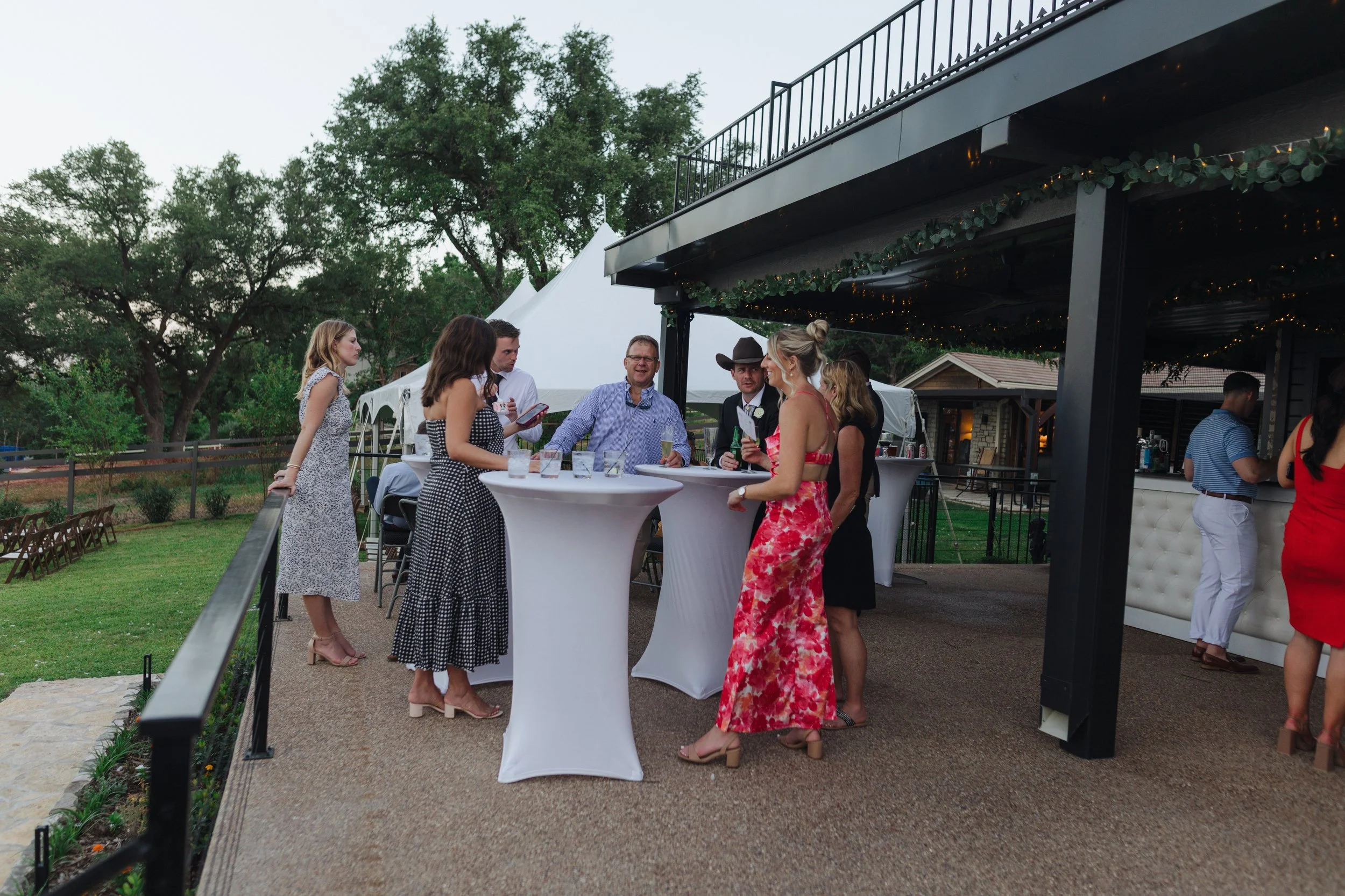 People socializing at a wedding reception outdoors, standing around high-top tables with drinks.