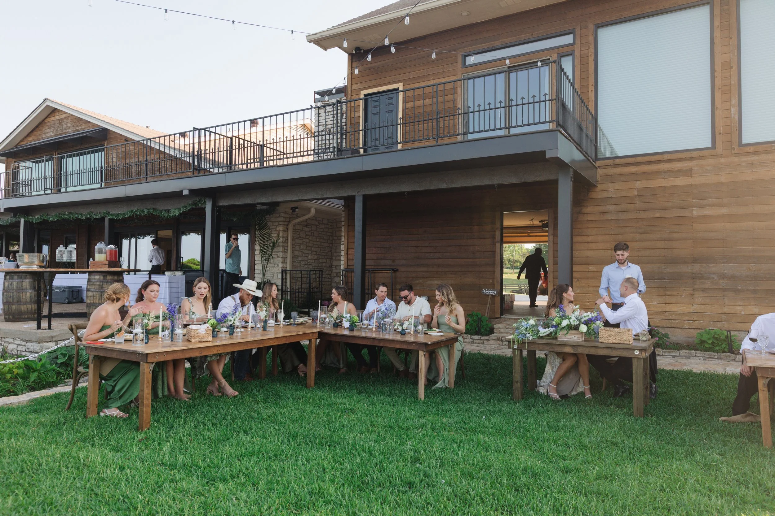 People sitting at long wooden tables outdoors during a celebration or gathering, with a modern wooden house in the background.