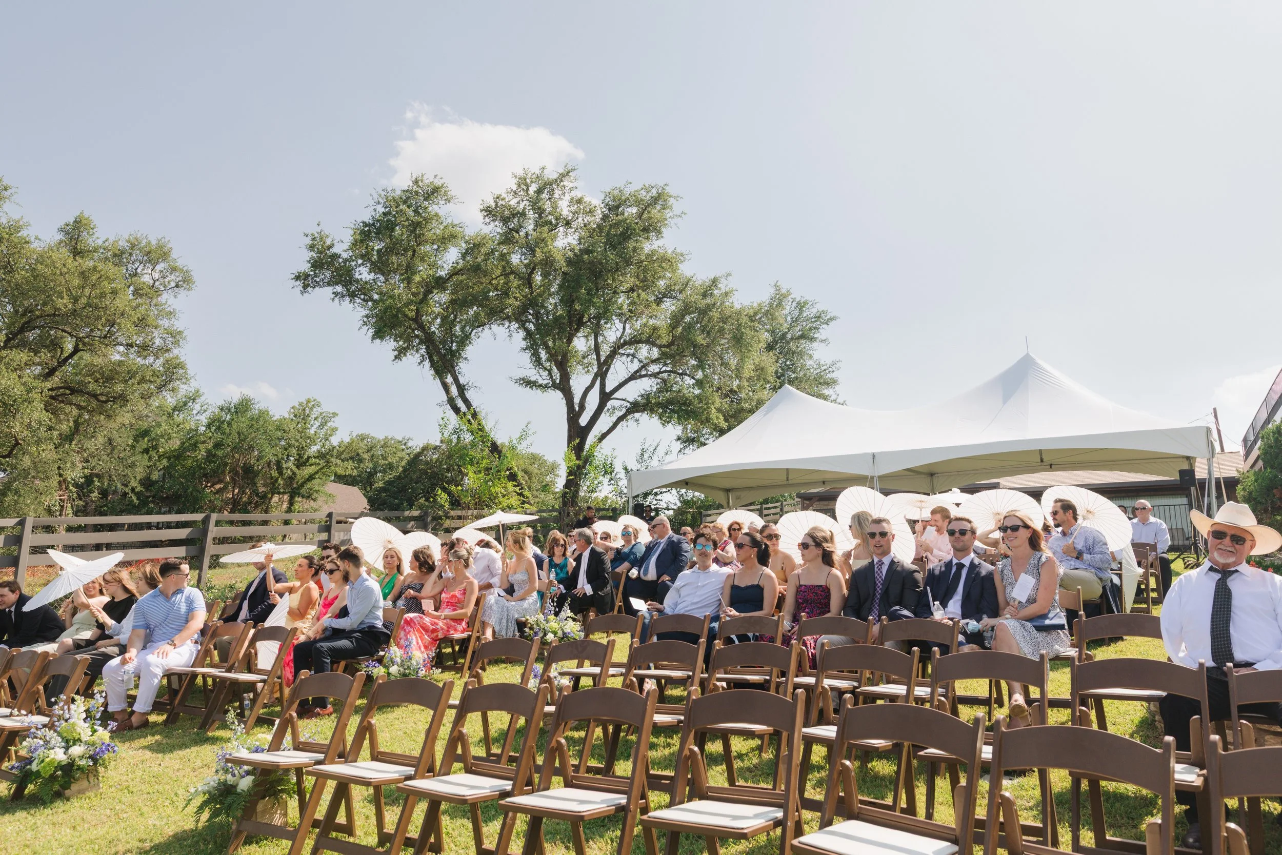 People seated outdoors at a wedding ceremony under white tents and umbrellas, with trees in the background on a sunny day.