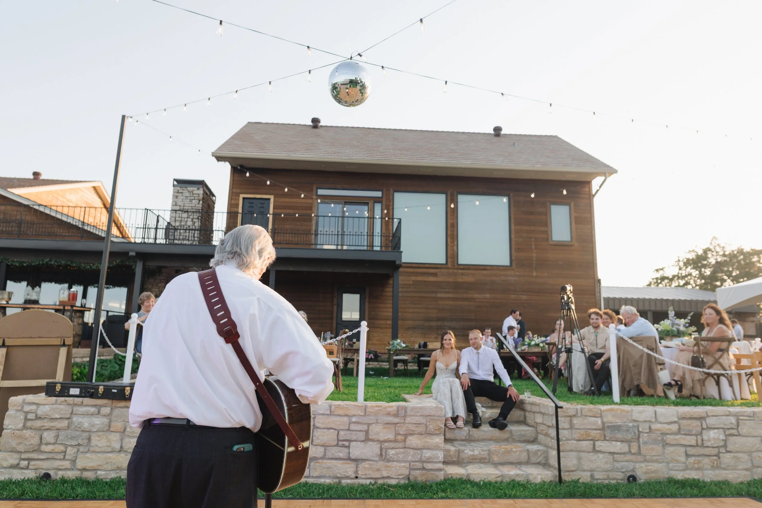 An outdoor wedding reception with a musician playing guitar in the foreground, and guests seated at tables and on steps, with a two-story house and string lights overhead.
