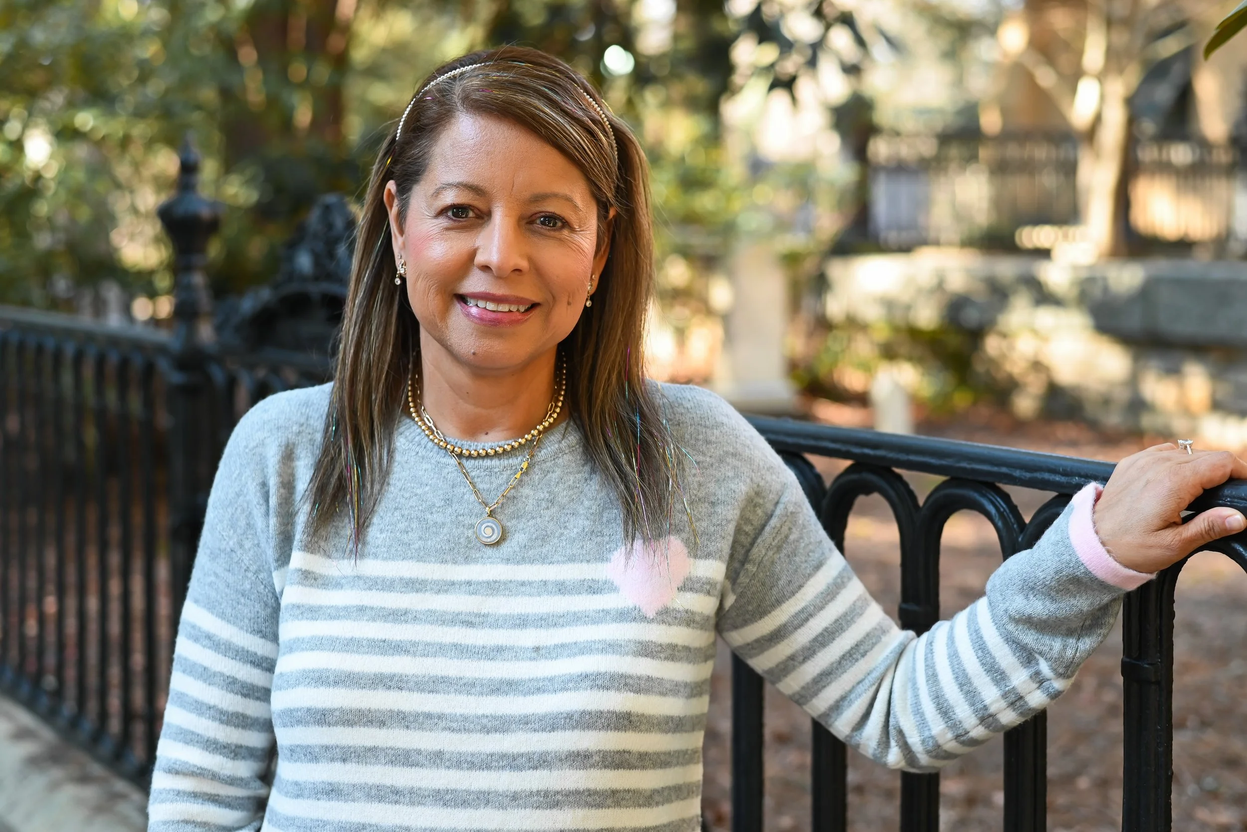 Smiling woman with shoulder-length brown hair, wearing a gray and white striped sweater, standing outdoors near a black iron fence with trees and greenery in the background.