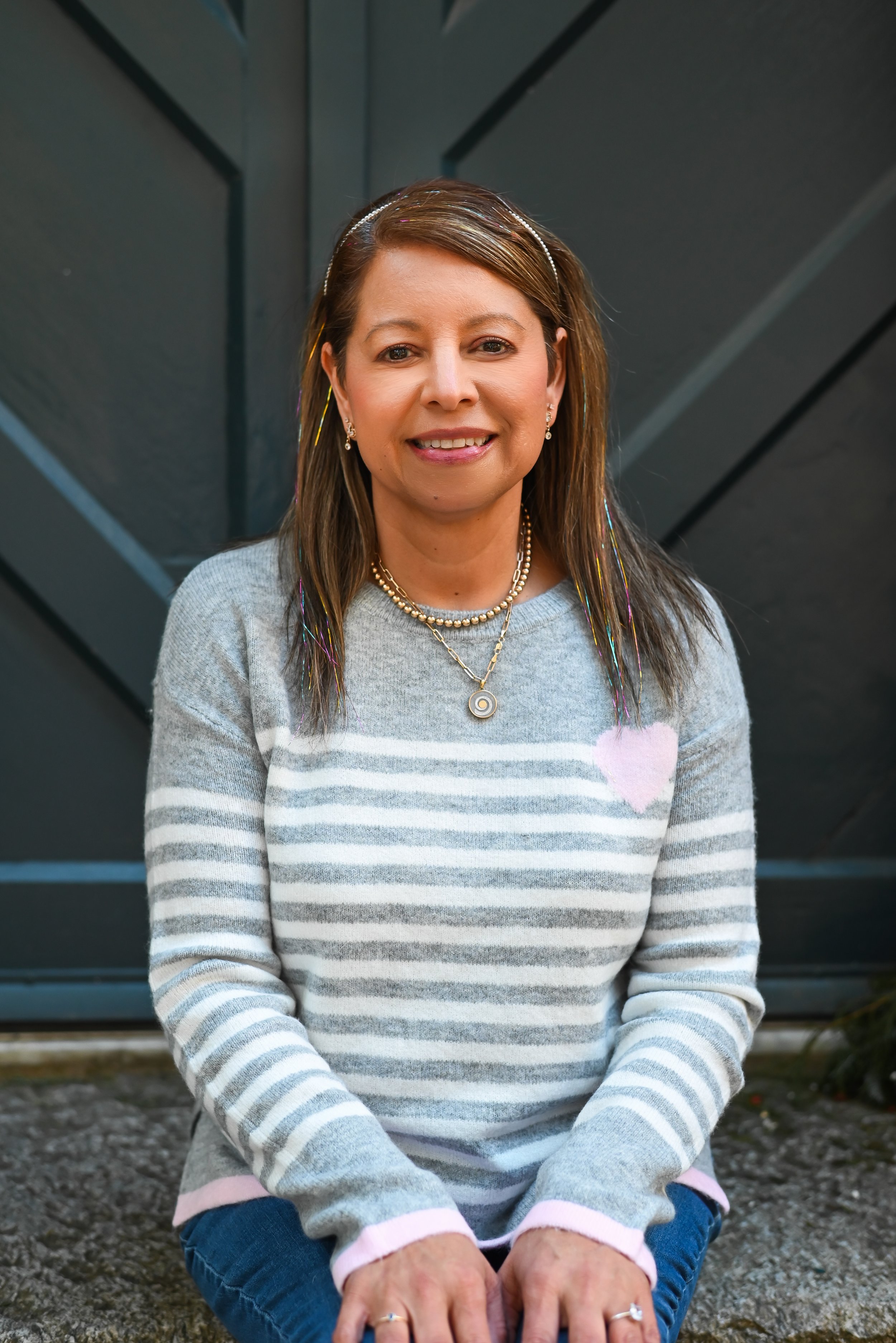 A woman sitting outdoors in front of a dark gray gate, smiling, wearing a gray striped sweater with pink accents, and layered necklaces.